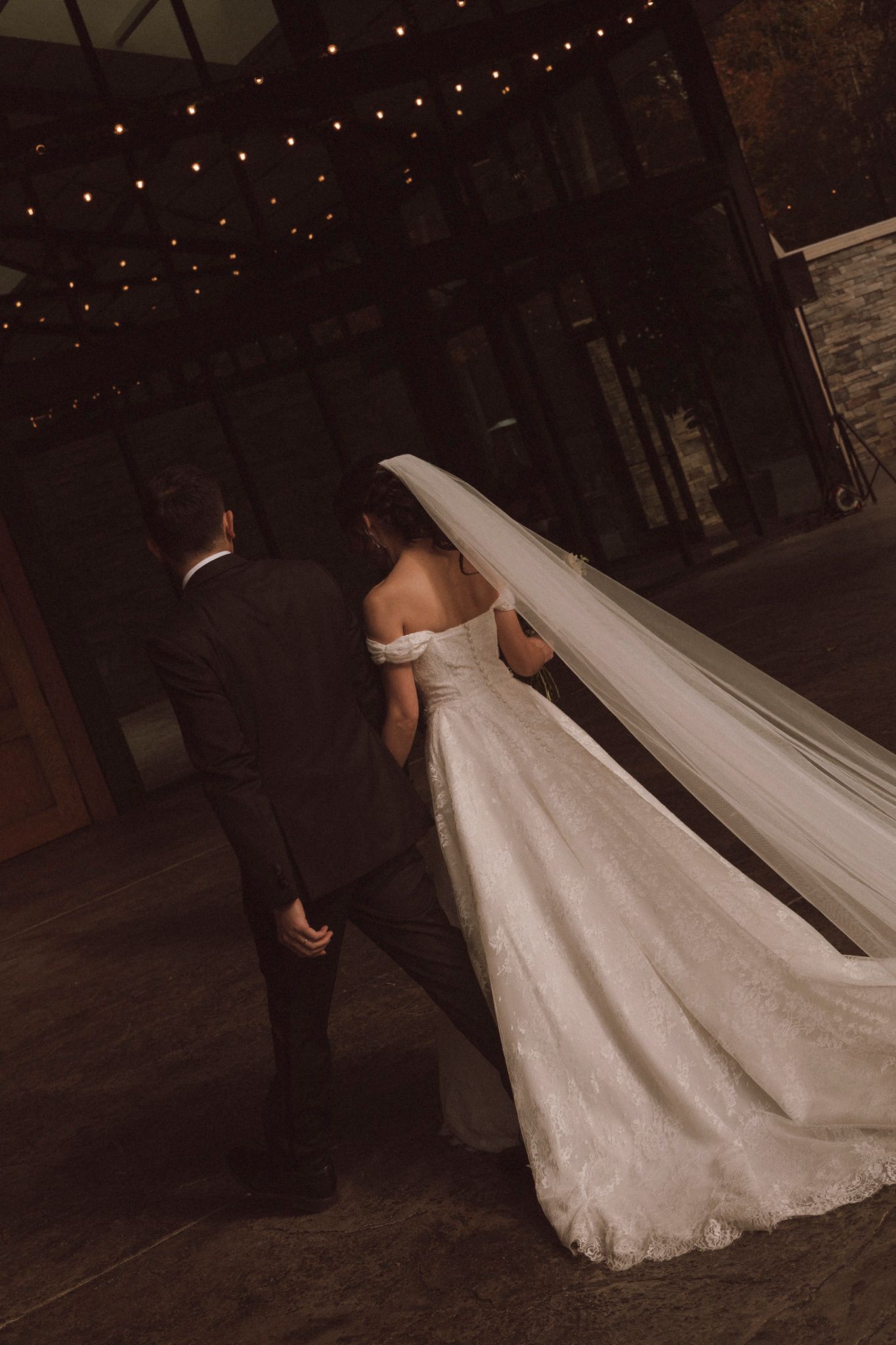A bride in a flowing white wedding gown with a long veil dances with her partner in a dimly lit venue.