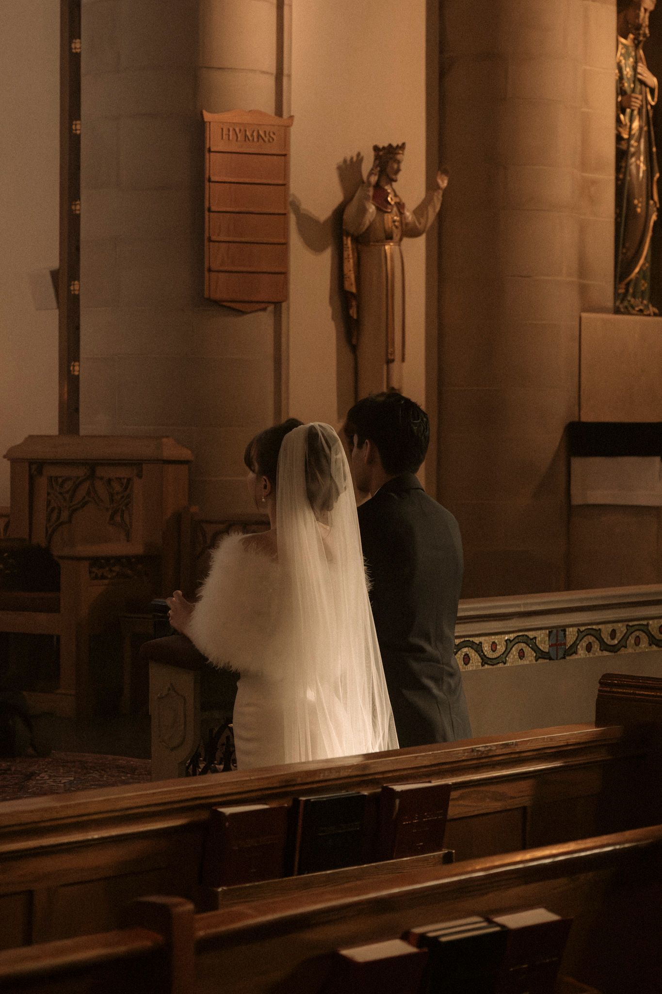 A bride and groom stand together in a dimly lit church with ornate columns and decorative details.