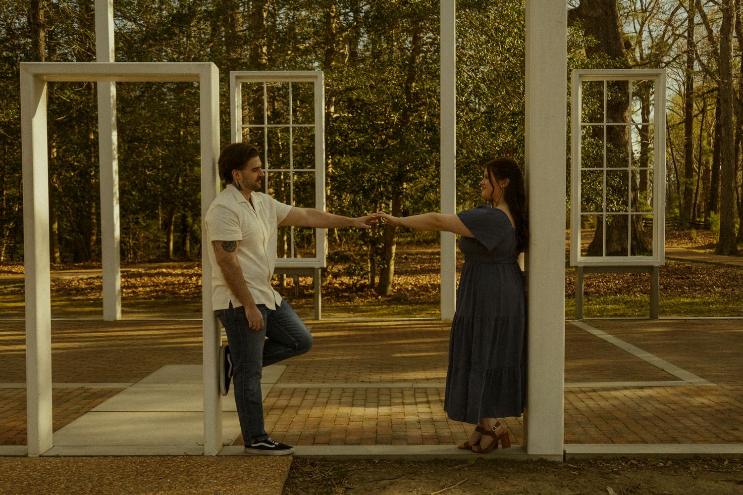 Couple holding hands between white window frames in an outdoor park setting with autumn trees in background.