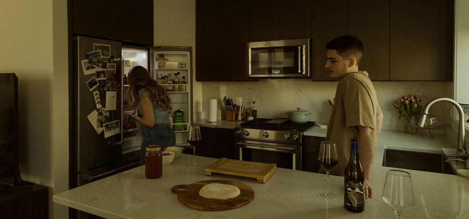 Two people in a modern kitchen, one browsing an open refrigerator while another stands at a marble counter with a wine bottle.