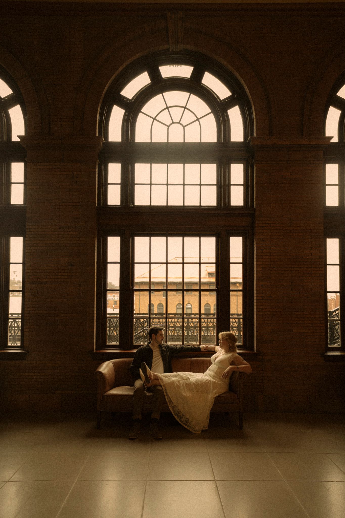 A couple in wedding attire rests on a couch by elegant arched windows in a grand historic building.