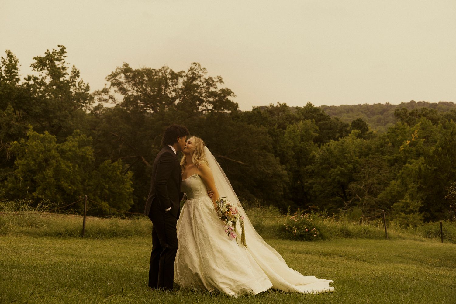 A bride and groom share a romantic kiss in a lush green field during golden hour at their outdoor wedding.