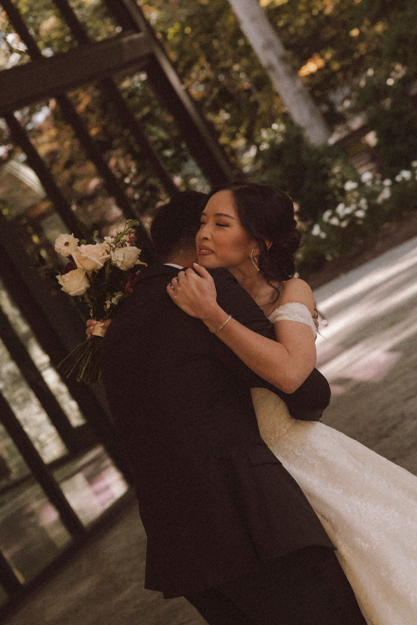 A romantic wedding couple embraces in a wooded outdoor setting while holding a white floral bouquet.
