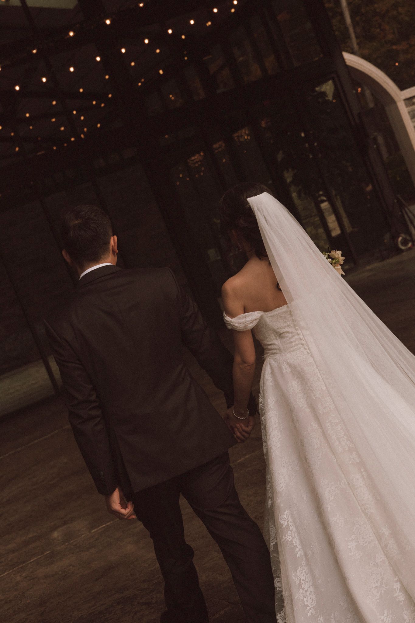A wedding couple walks together under twinkling lights during their evening celebration.