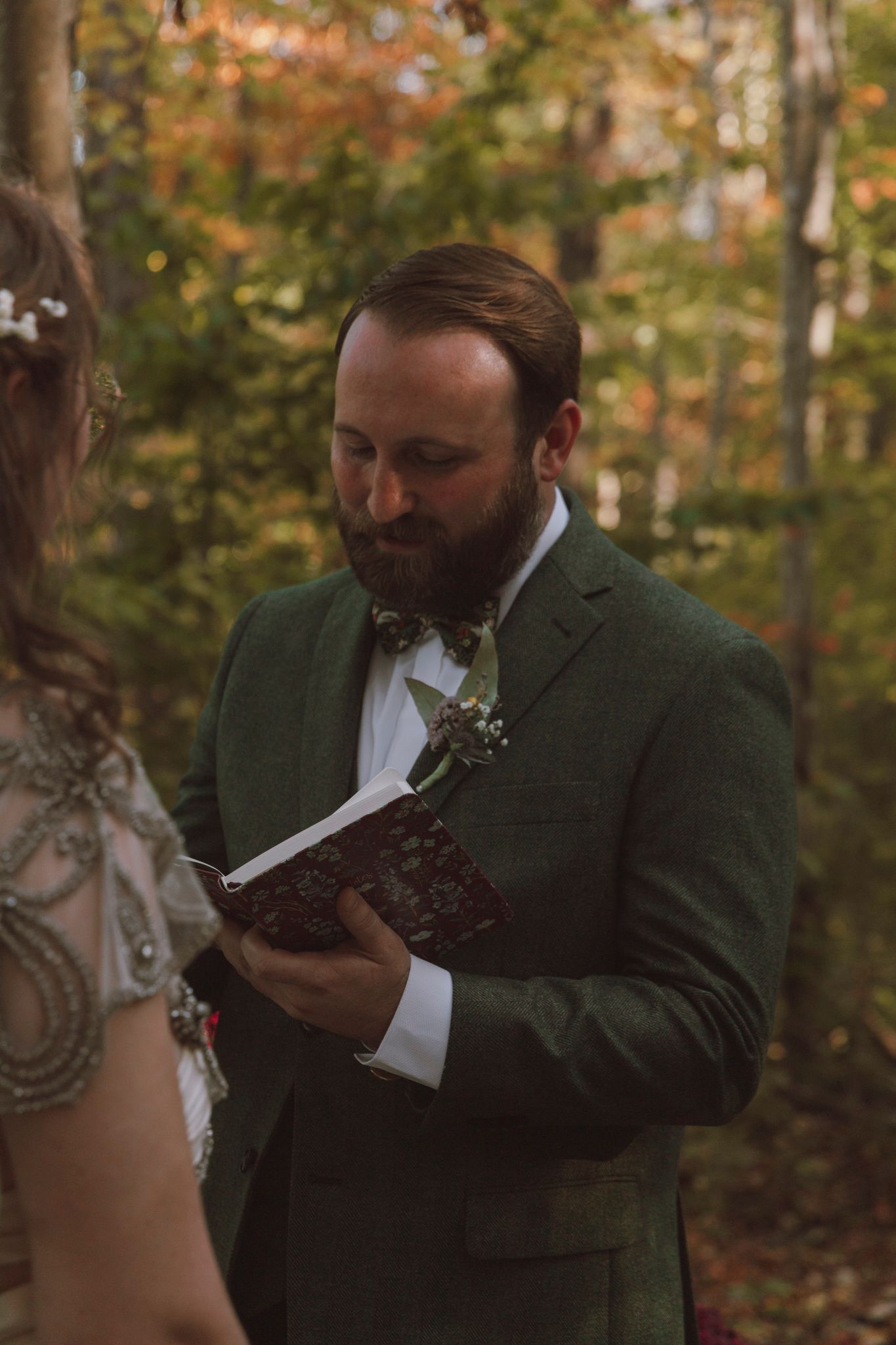 A groom in a green suit jacket reads from a book during an autumn outdoor wedding ceremony.