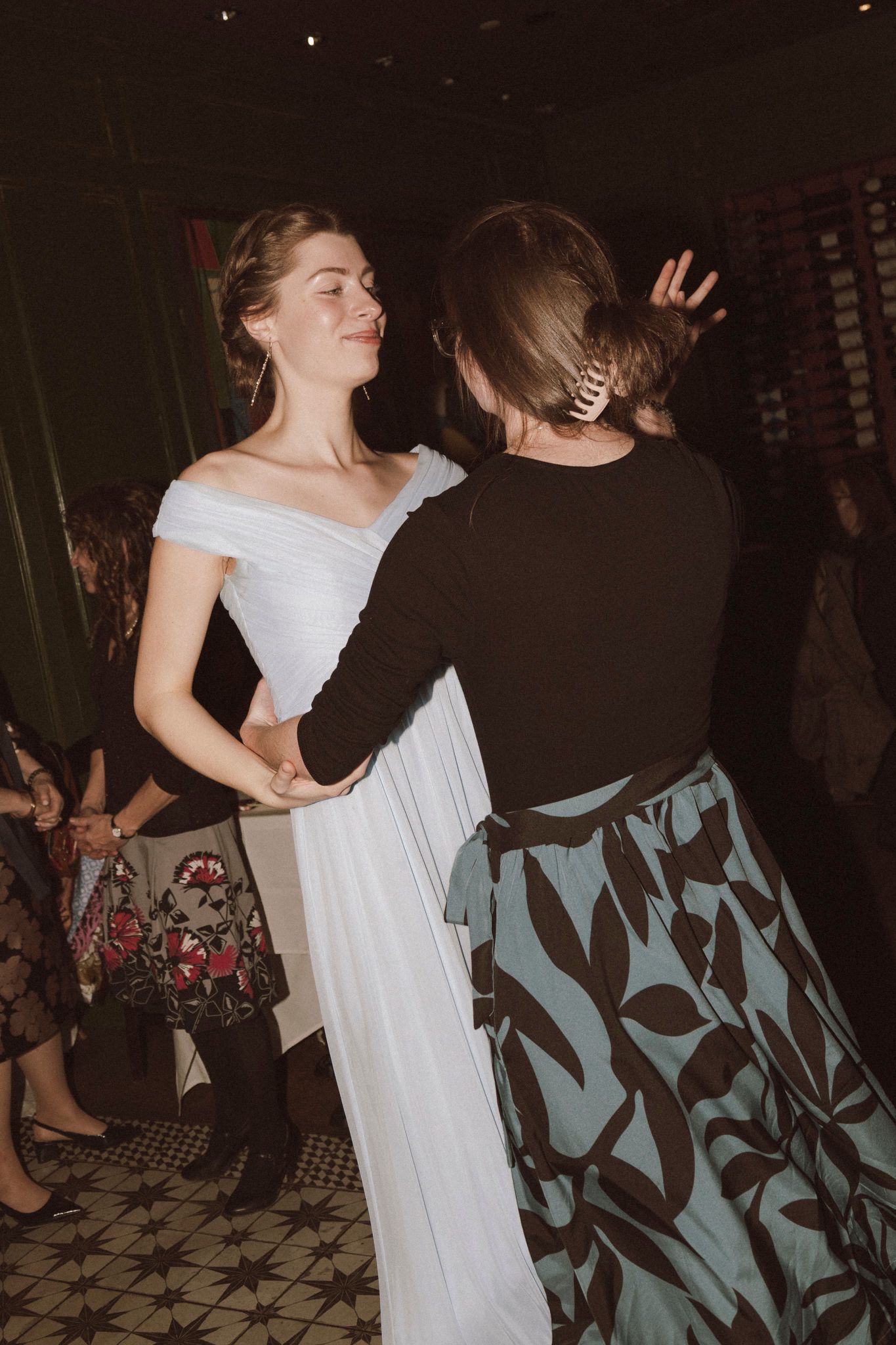 Bride in white off-shoulder gown dancing joyfully with female guest at wedding reception.