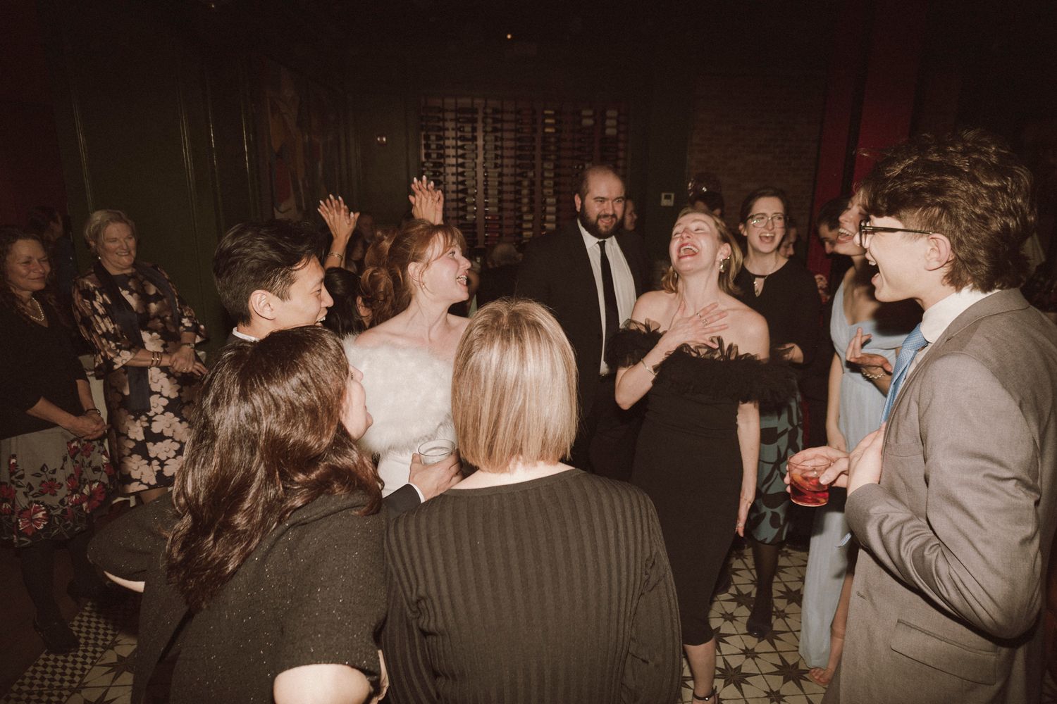 Wedding guests laugh and celebrate together with bride in white dress at indoor reception.