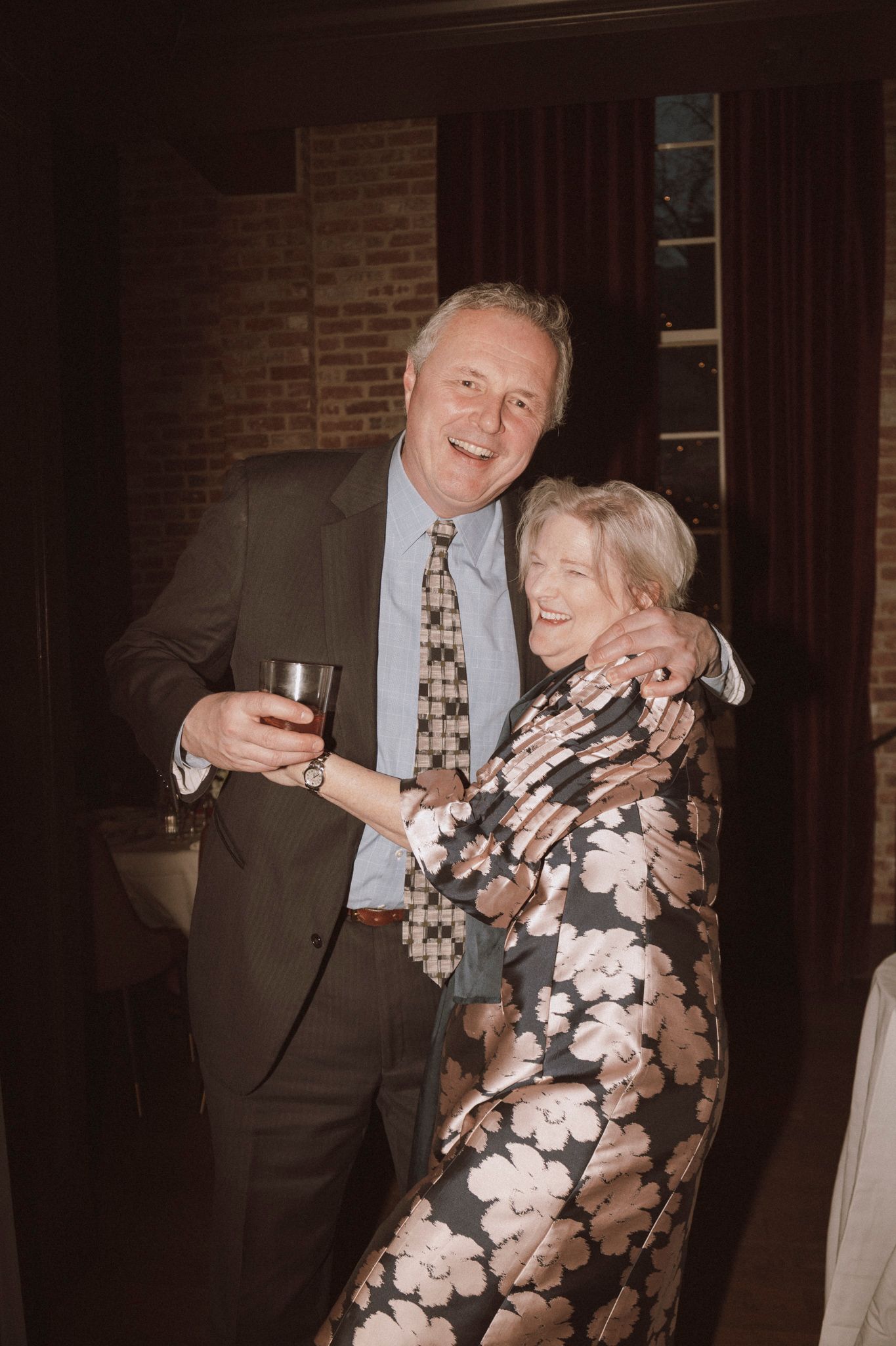 A smiling man in a suit holding a drink embraces a woman in a floral dress at a social gathering.