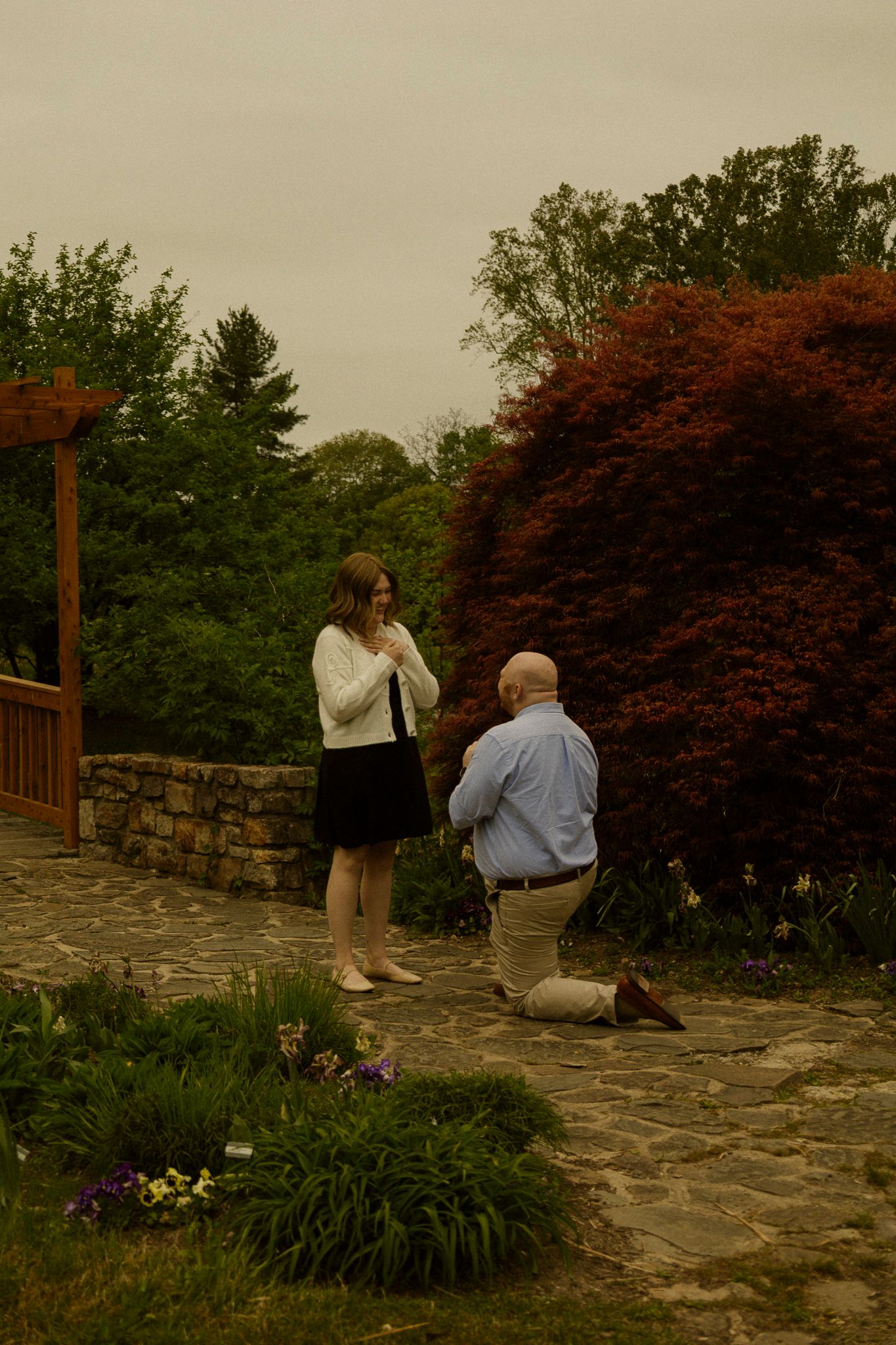 Man kneeling on stone path proposing to woman in garden surrounded by lush green and red foliage.