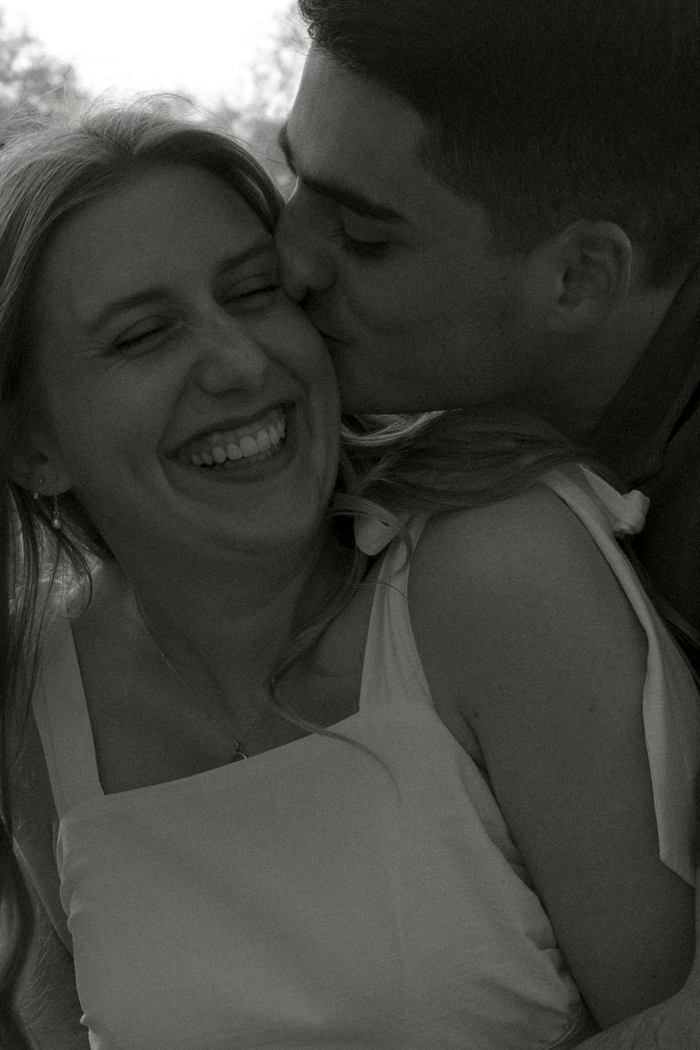 Black and white photo of a smiling woman being kissed on the cheek by a man in a romantic outdoor setting.