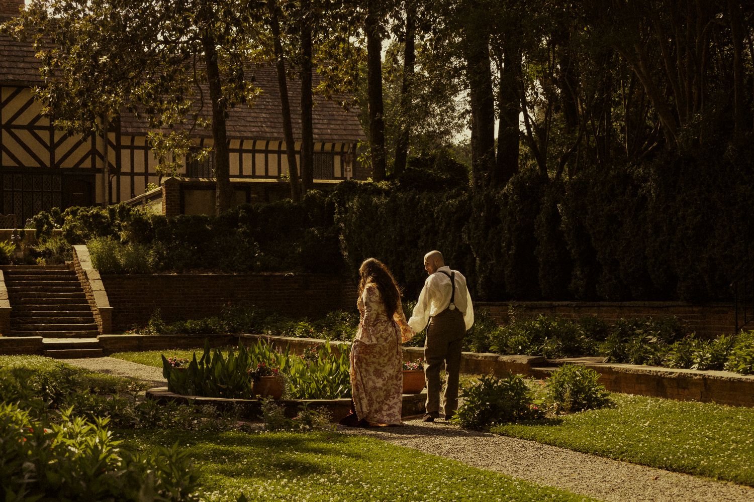Couple walking hand in hand through a lush garden beside a Tudor-style building at golden hour.
