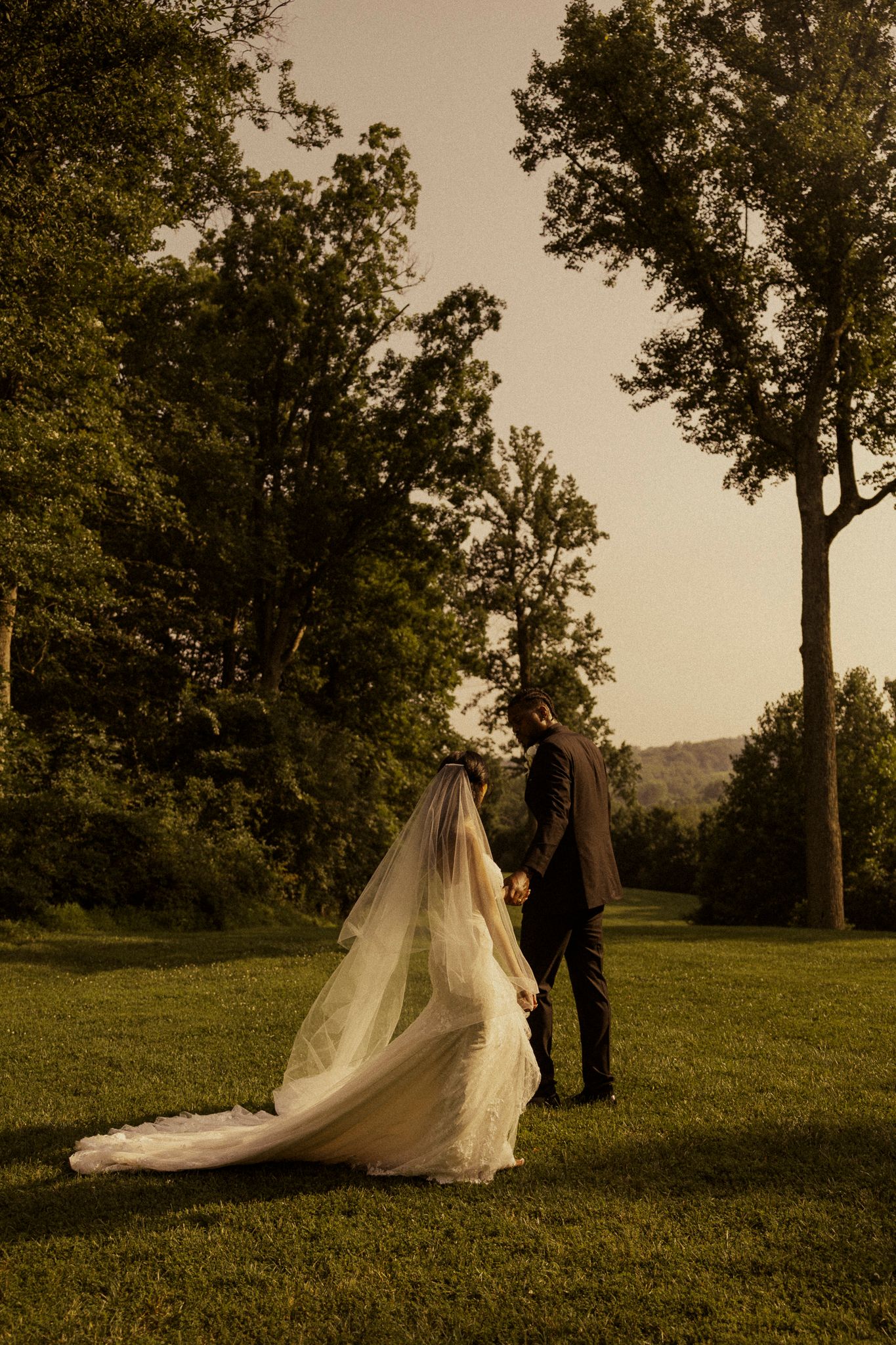Bride in flowing white gown and veil holds hands with groom in suit on green lawn at golden hour.