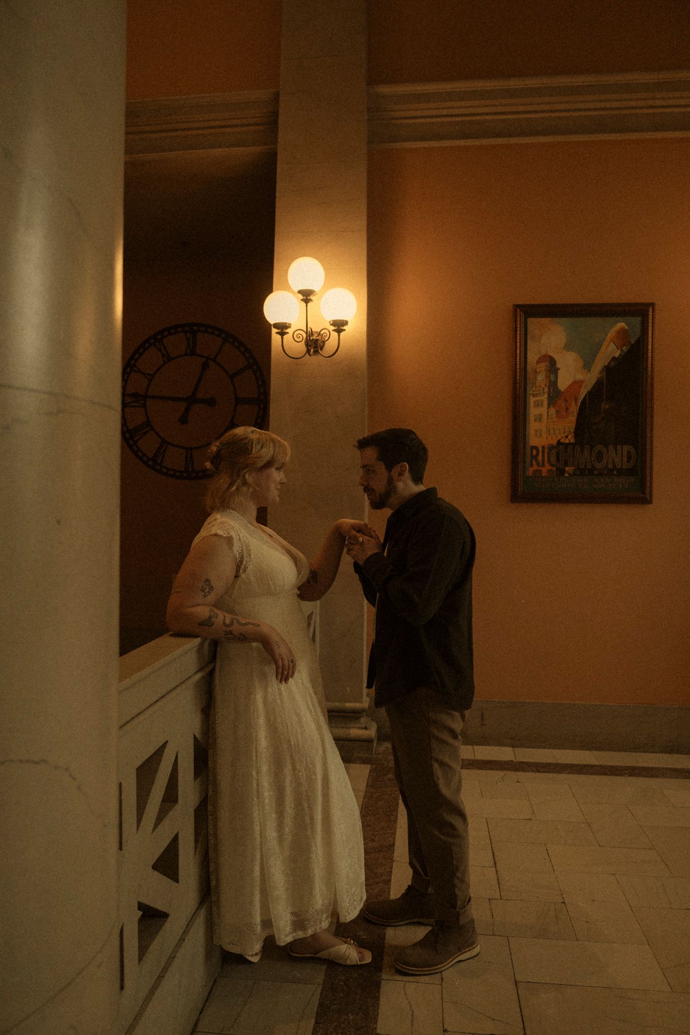 A couple in formal attire stand together in an elegant hallway with columns and wall sconces.