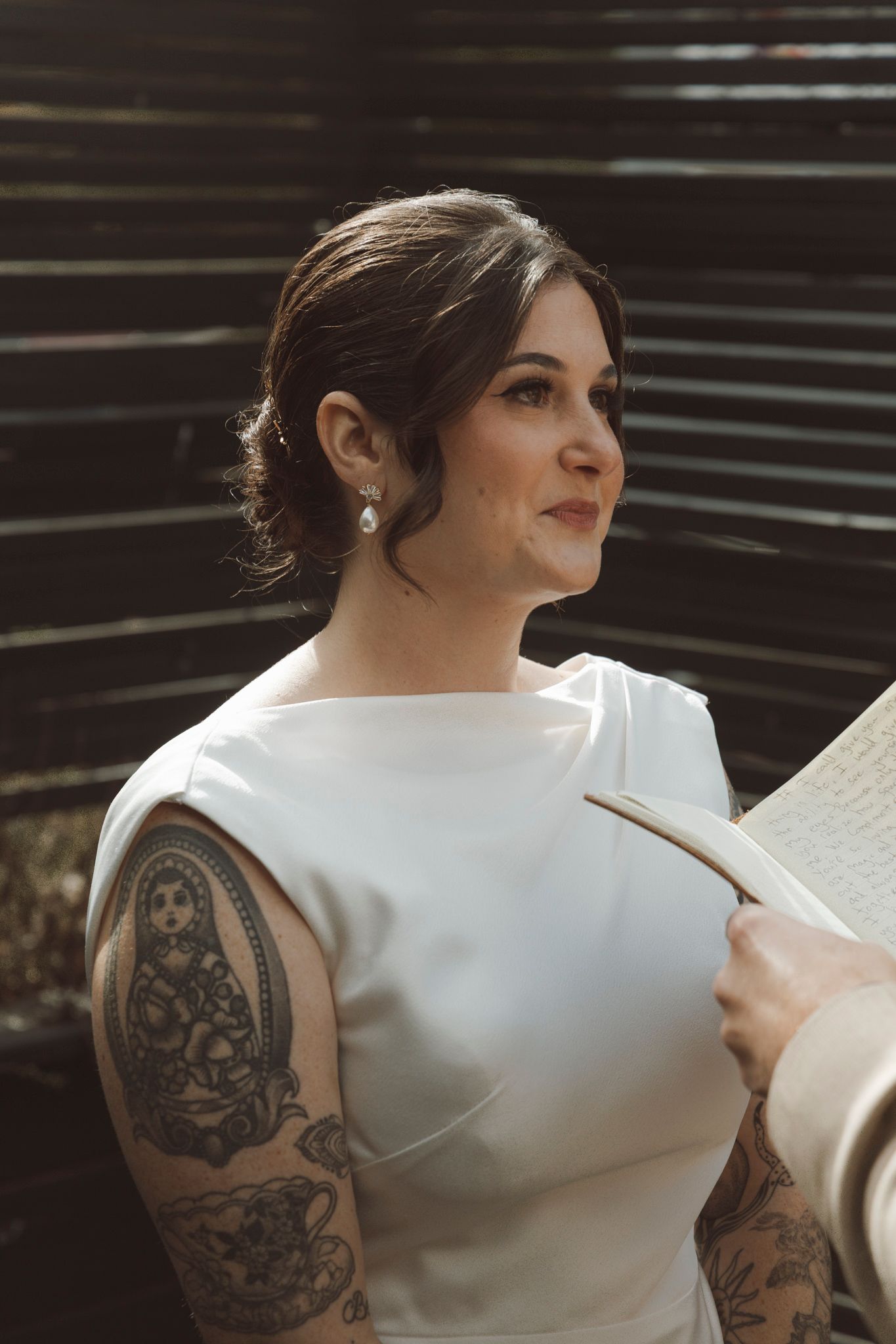 Person in white sleeveless dress with ornate tattoo on upper arm sits in elegant pose.