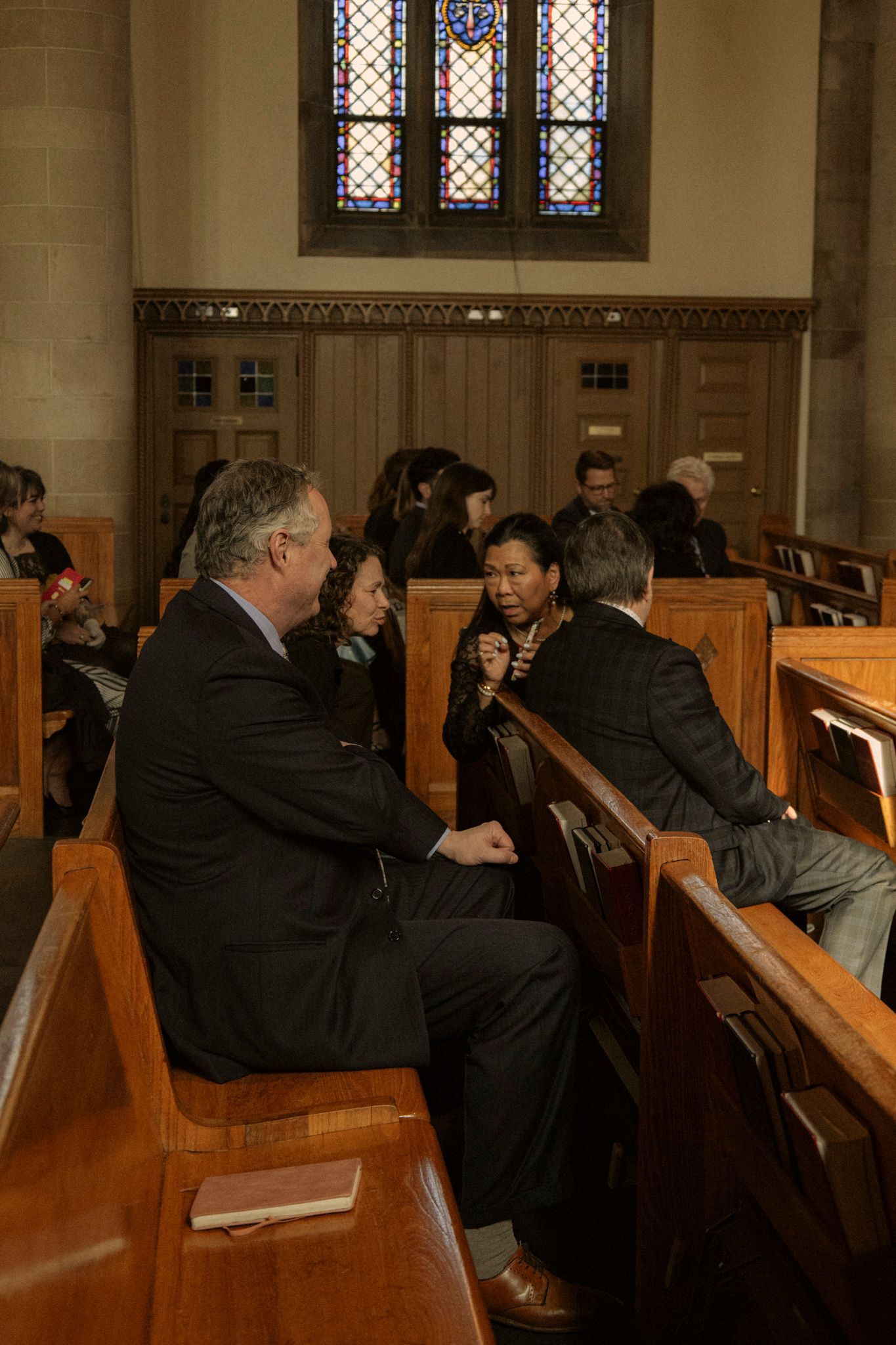 People conversing in wooden church pews beneath a stained glass window during a religious gathering.