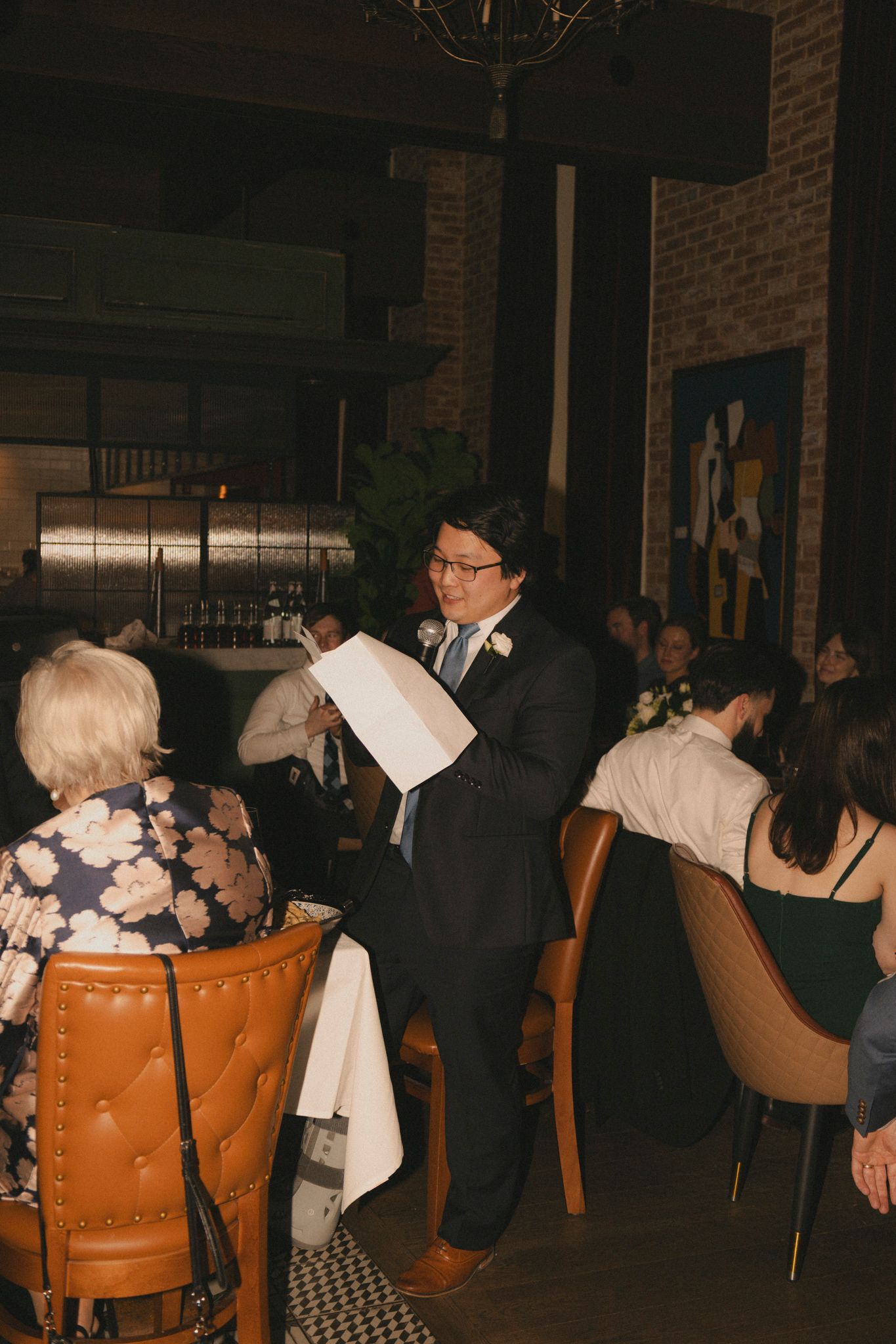 Groom in dark suit reads a speech at wedding reception in a dimly lit restaurant with brick walls.