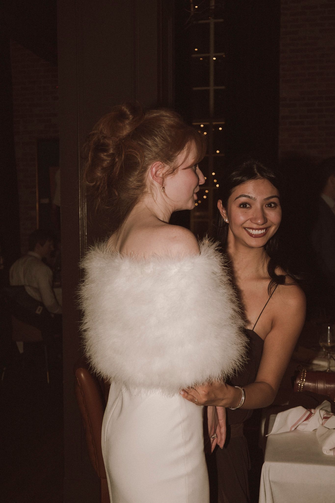 Bride in white dress with feather wrap shares a joyful moment with smiling bridesmaid at reception.