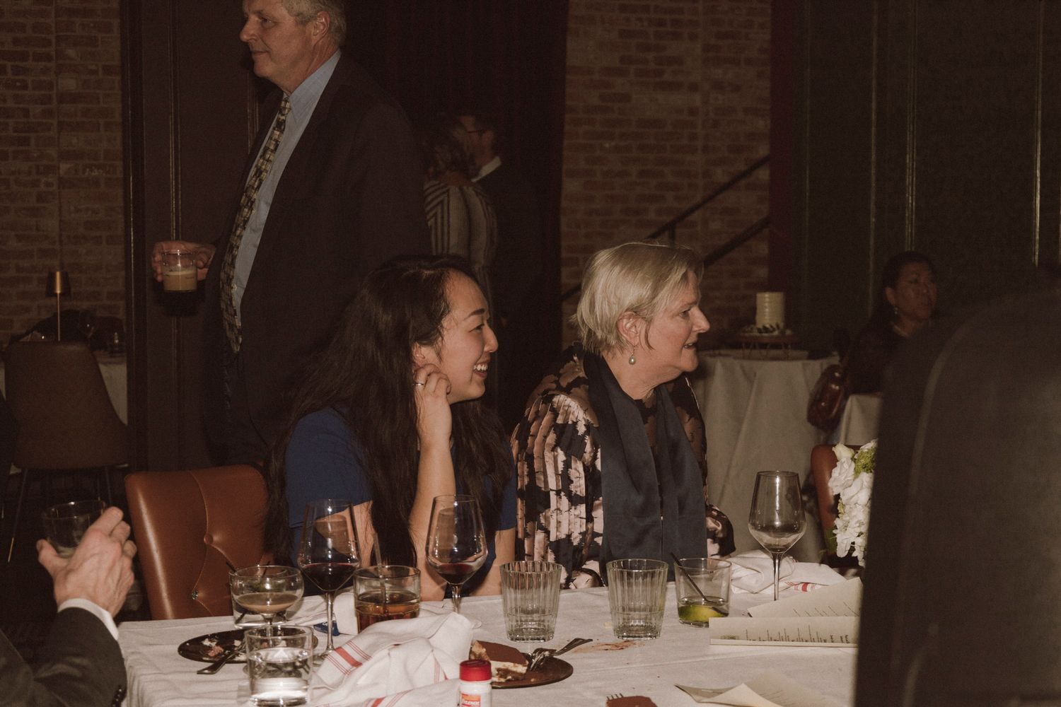 Guests seated at wedding reception dinner table with wine glasses in brick-walled venue.