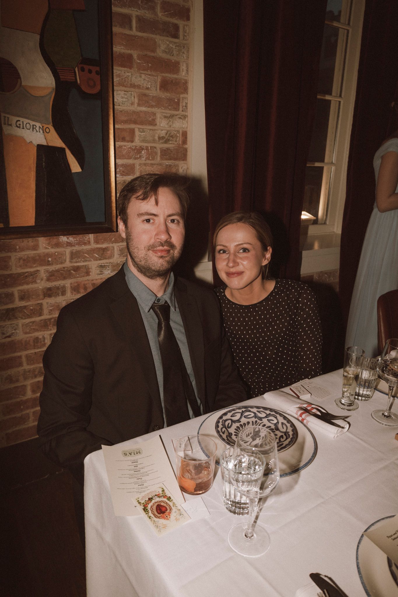 Couple smiling together at a restaurant dinner table with white tablecloth, brick wall and artwork in background.
