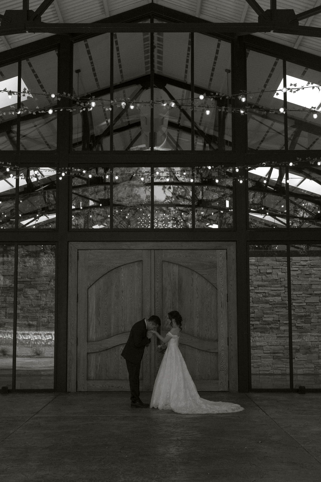 A romantic black and white wedding photo of a couple silhouetted against grand wooden doors in a rustic venue.