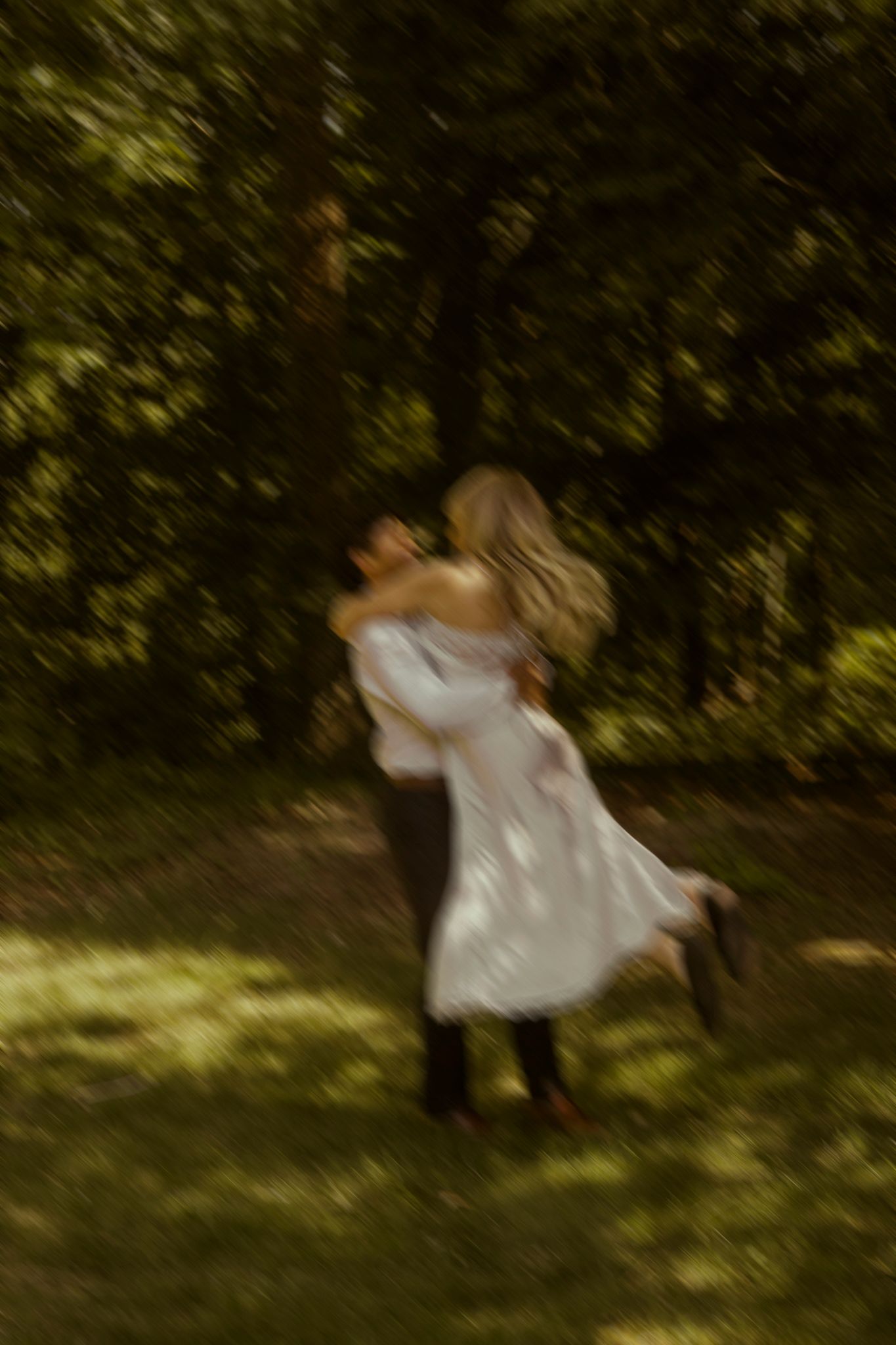 Blurred motion photo of a couple embracing and dancing in a sunlit forest clearing, woman in white dress.