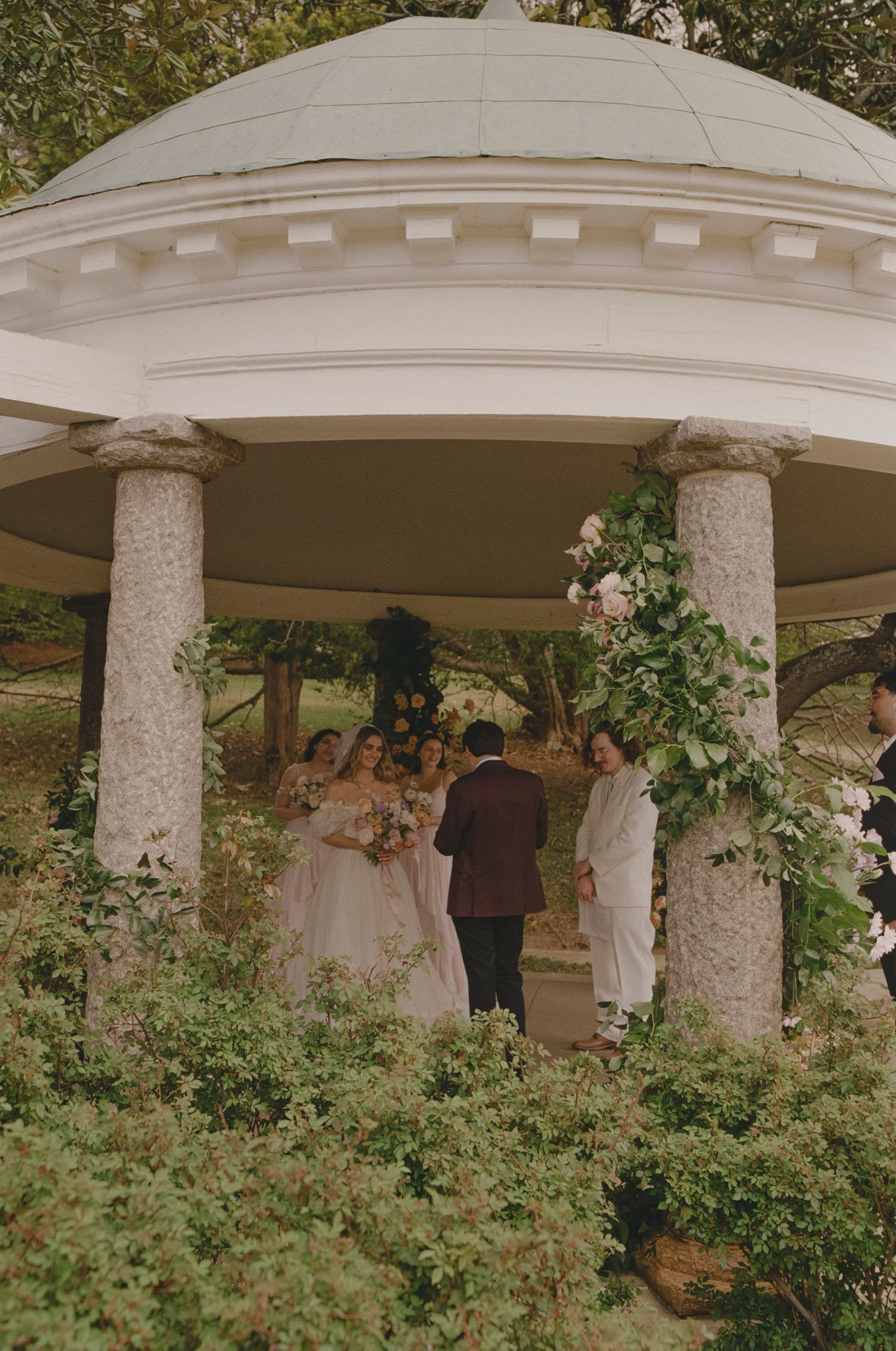 A romantic outdoor wedding ceremony takes place under a white columned gazebo surrounded by lush greenery.