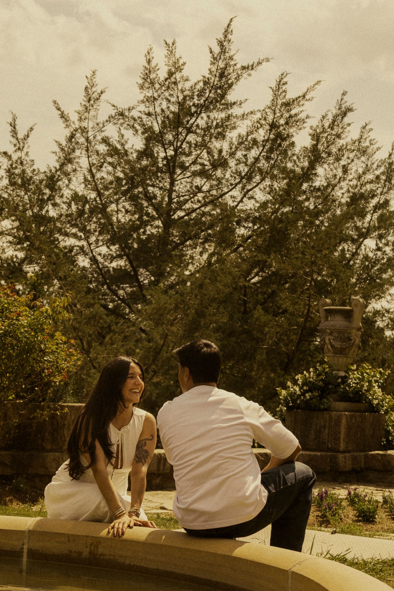 A smiling couple sits together outdoors near a fountain, surrounded by lush green trees on a warm day.