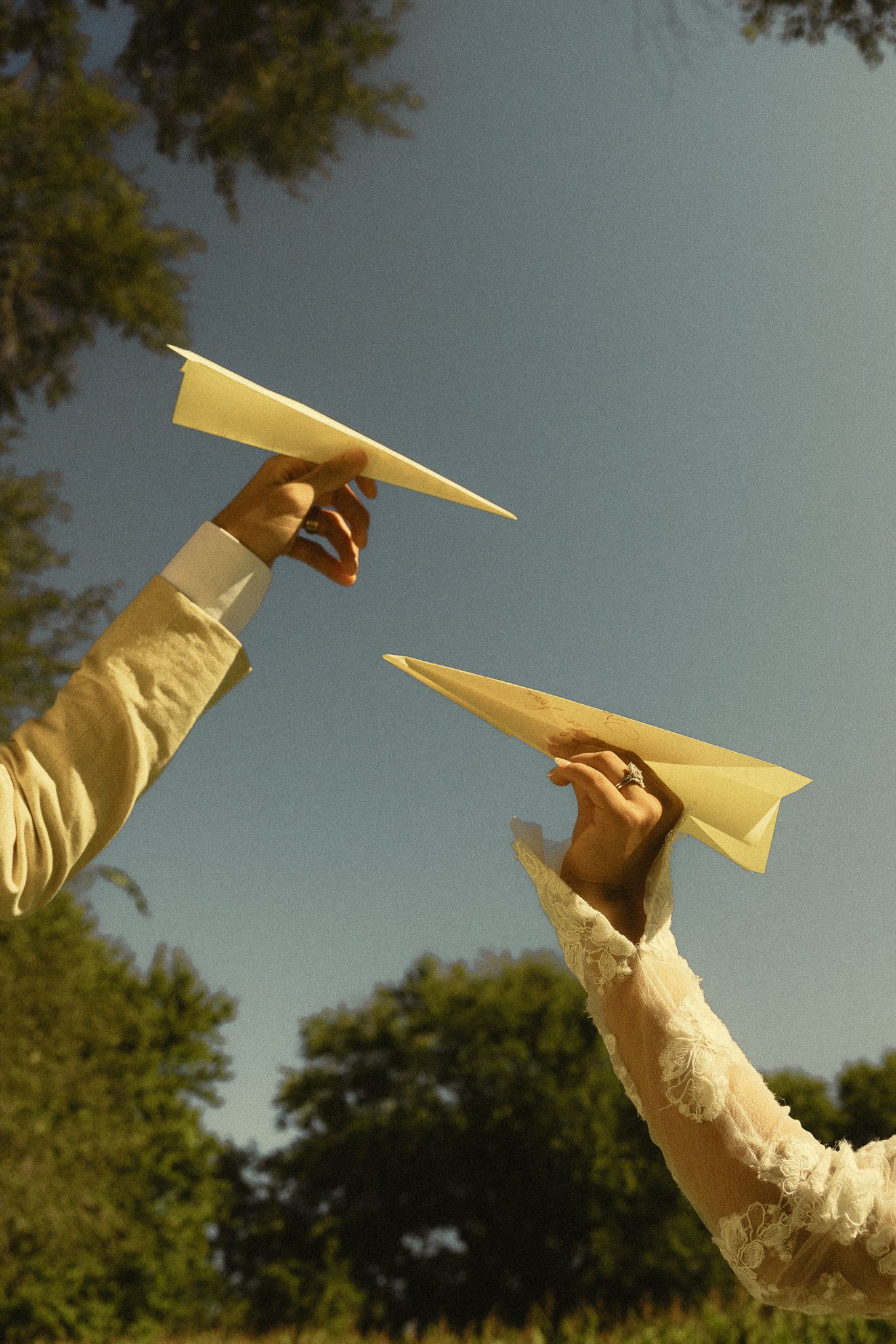 Bride and groom holding paper airplanes up to blue sky at outdoor wedding celebration.