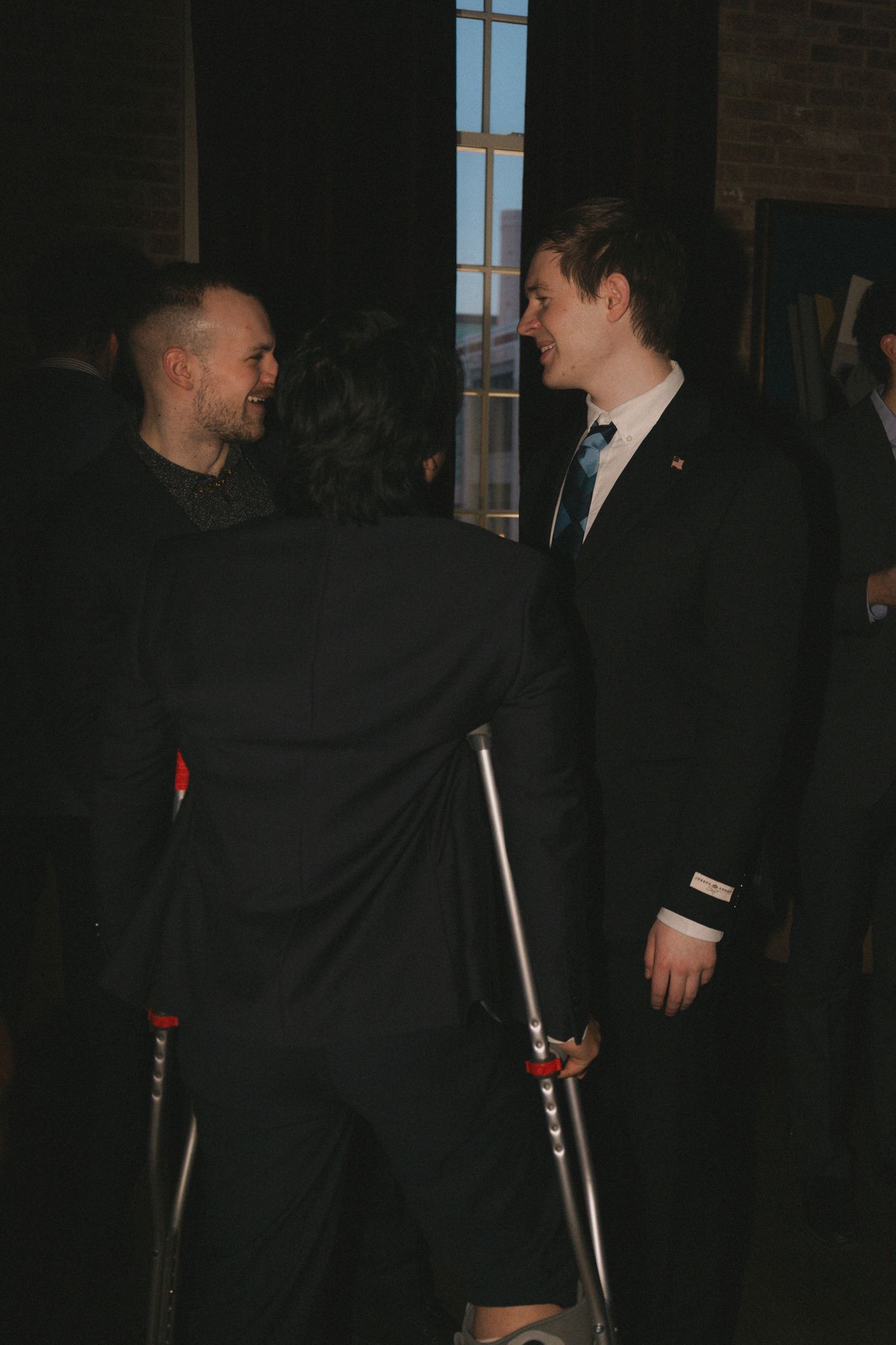 Two men in suits converse at a formal event, one using forearm crutches, in a dimly lit venue.