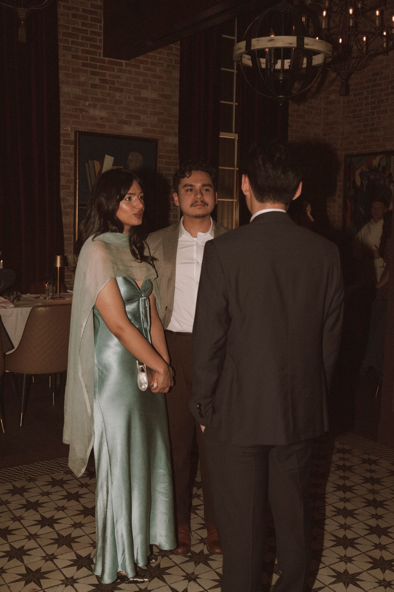 Wedding guest in elegant green satin cape dress converses with guests on patterned floor in brick-walled venue.