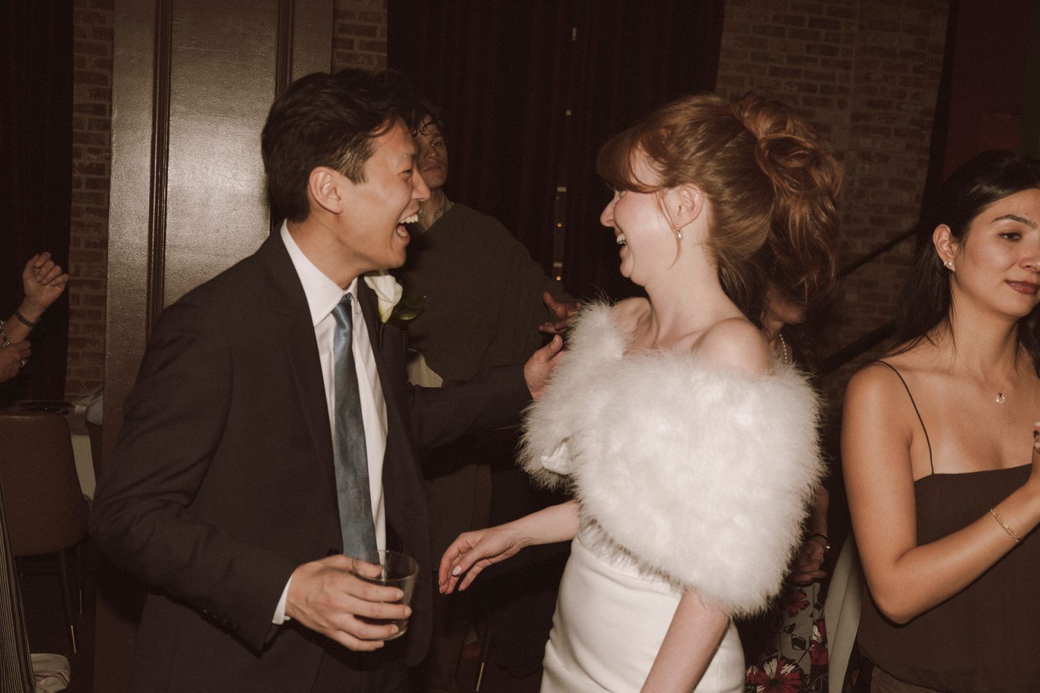 Bride in white gown with feather wrap laughs joyfully with a male guest at wedding reception.