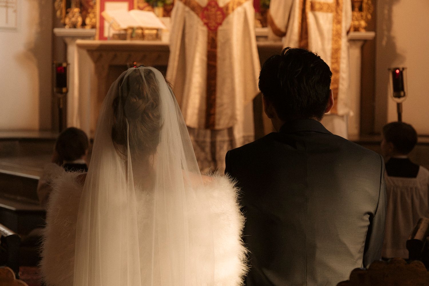 Bride with veil and groom sit before priest at ornate golden altar during Catholic wedding Mass.