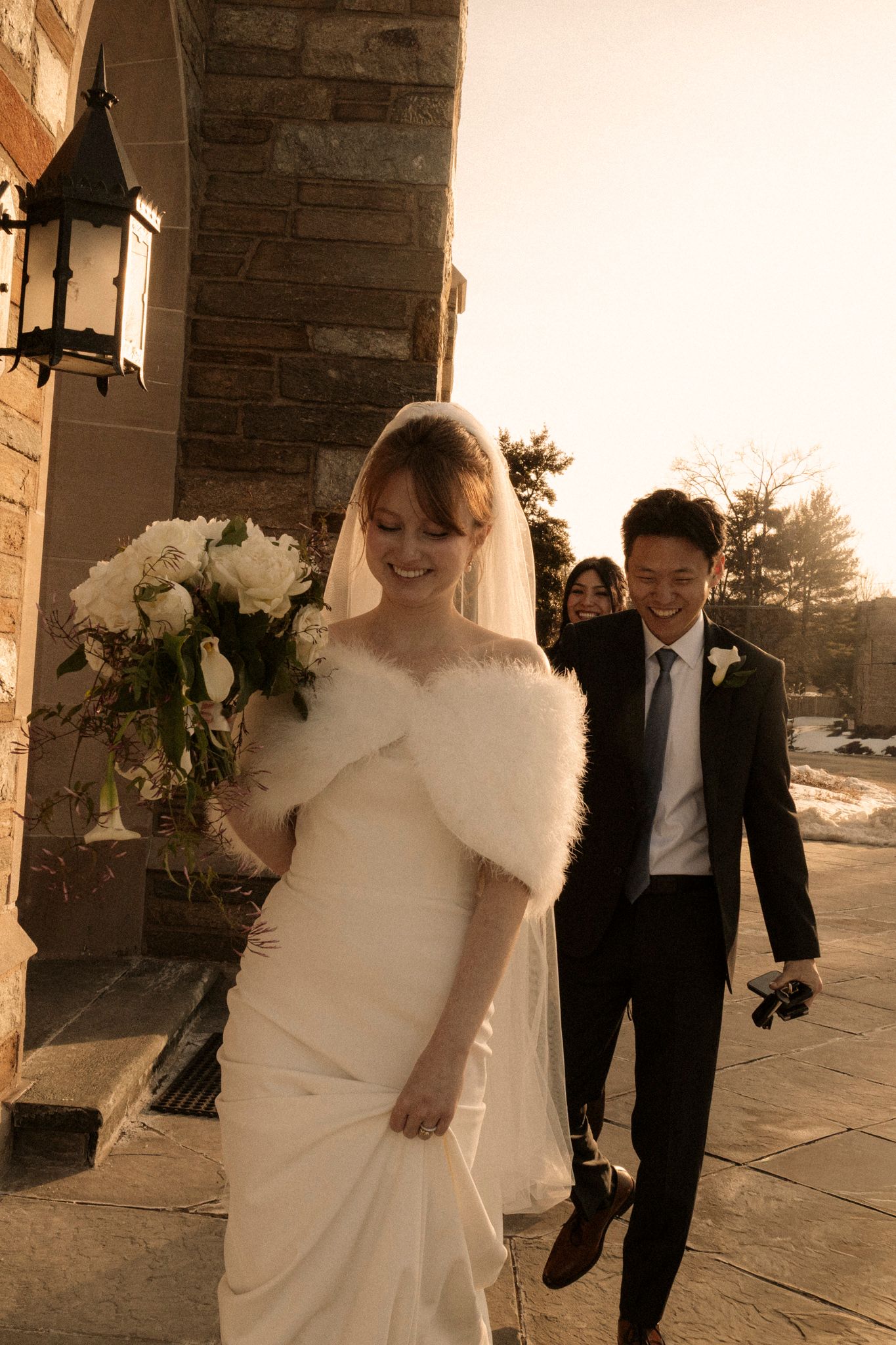 Smiling bride in white gown, fur stole, and veil holds bouquet while walking with groom in black suit outside stone church.