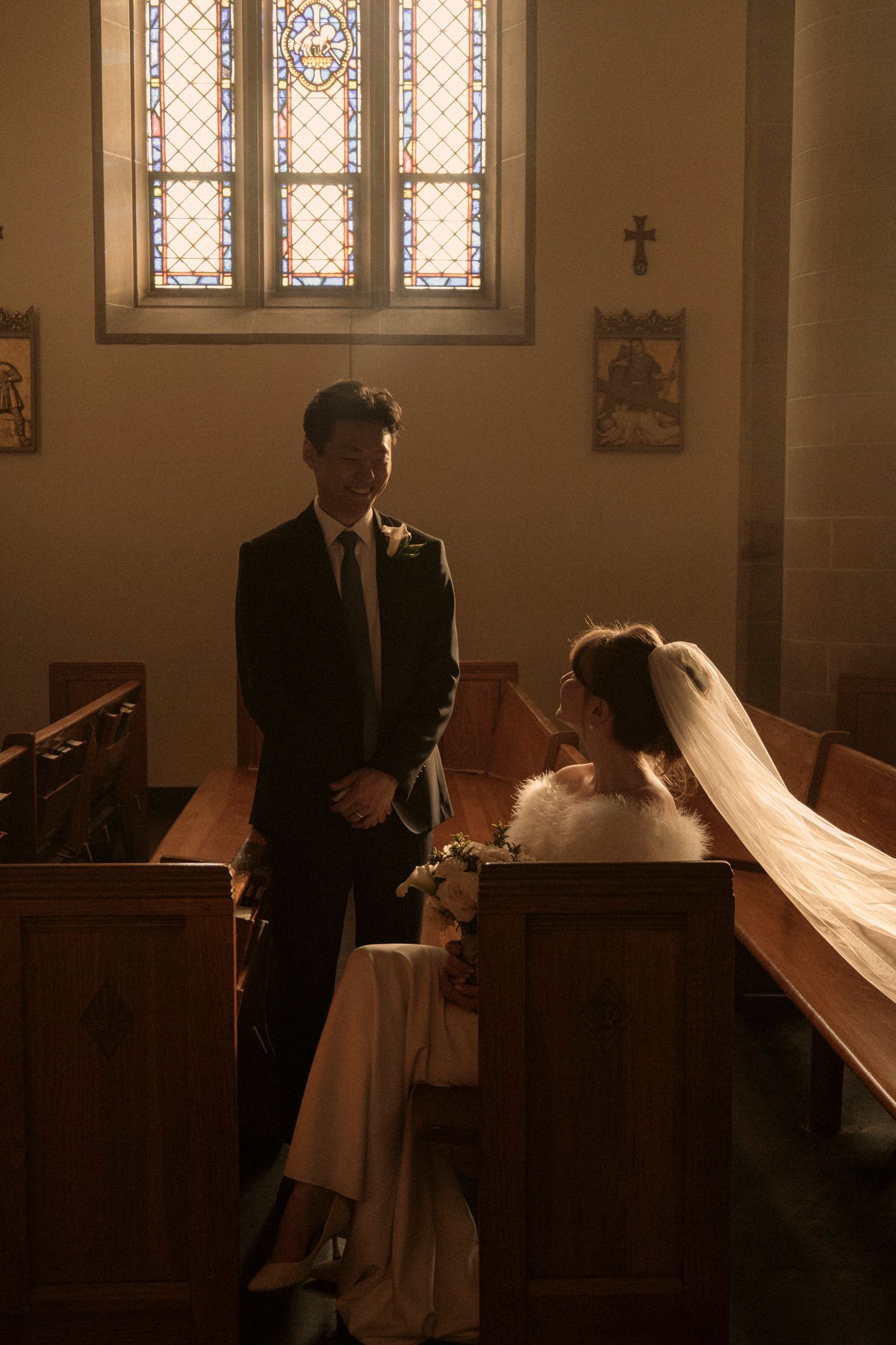 Bride and groom share a tender moment inside a sunlit church with stained glass windows and wooden pews.
