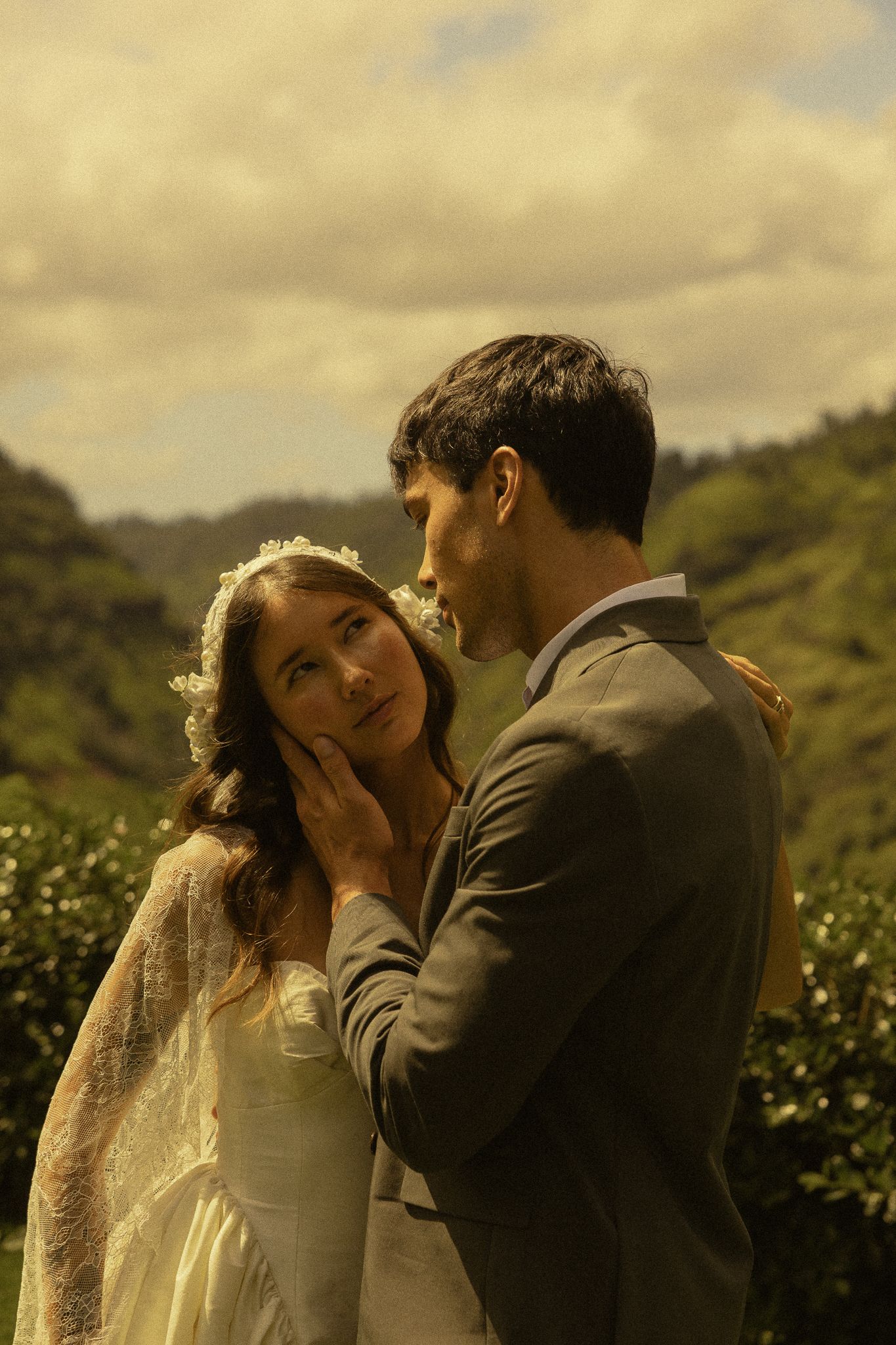 A groom tenderly cups his bride's face outdoors, with lush green hills and cloudy sky behind them.