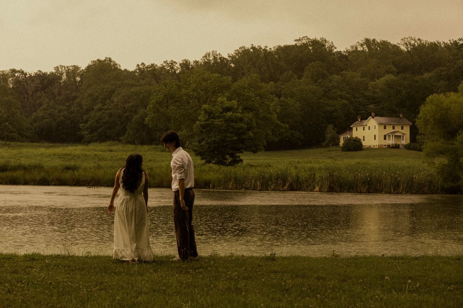Couple standing by a serene lake at dusk with lush green trees and a white farmhouse in the background.