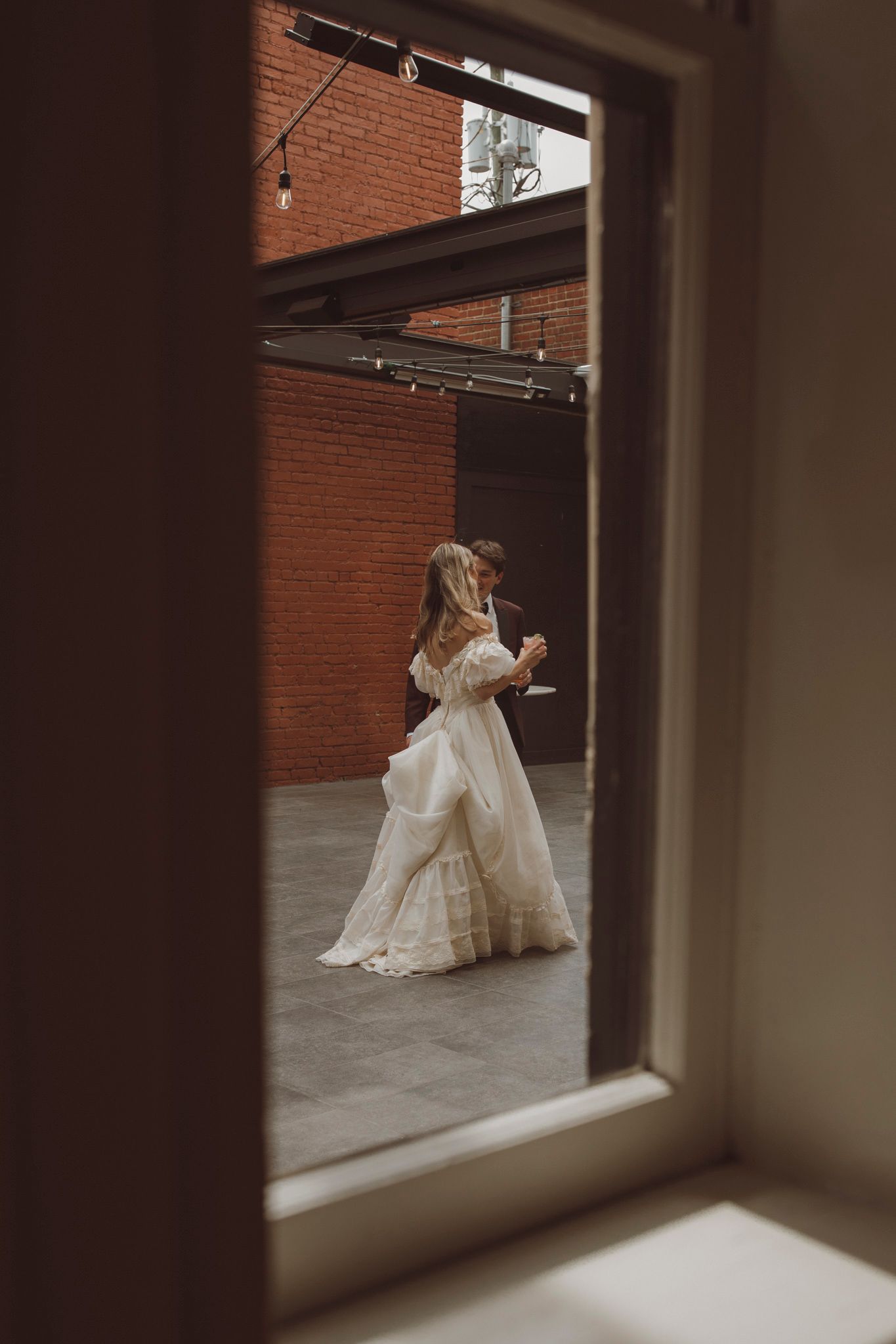 Through a window frame, a bride in a flowing white wedding dress twirls gracefully in an industrial space with brick walls.