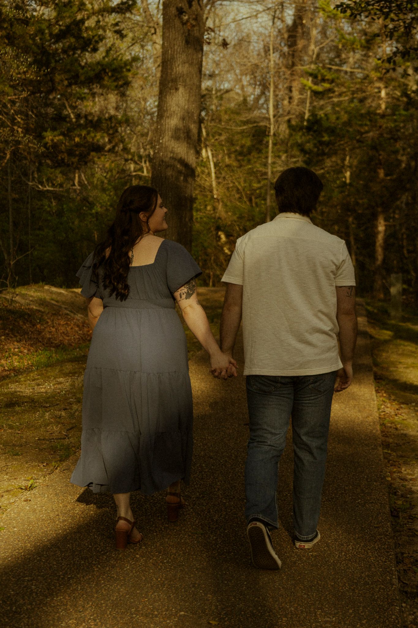Couple holding hands walking down a sunlit forest path, woman in gray dress and man in white shirt and jeans.