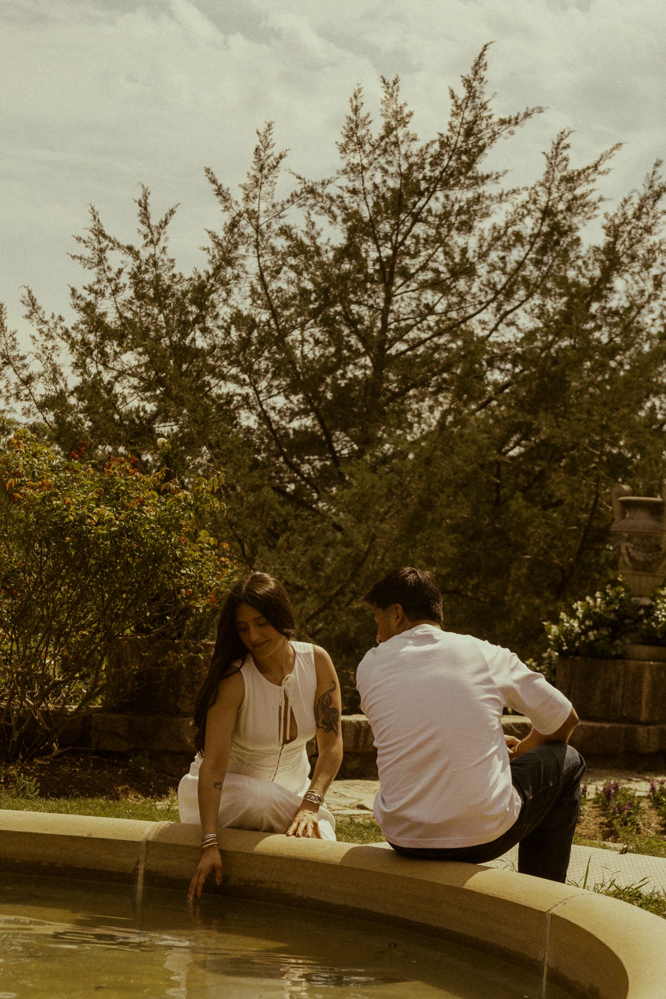 Couple sitting by a garden fountain, woman in white dress touching water, man in white shirt, lush green trees behind.