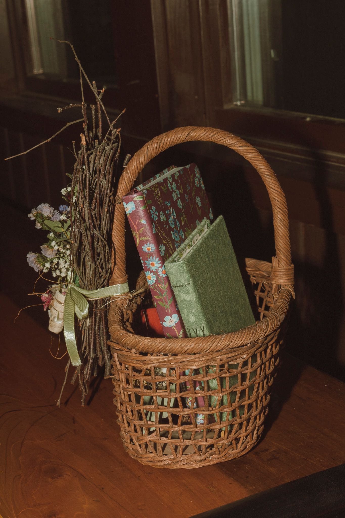 Vintage woven basket with books and dried floral arrangement on wooden surface in moody lighting.