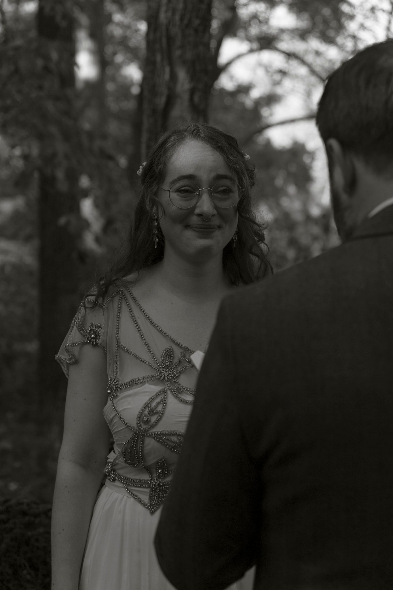 A black and white photo shows someone wearing glasses and an embellished dress standing in a wooded setting.