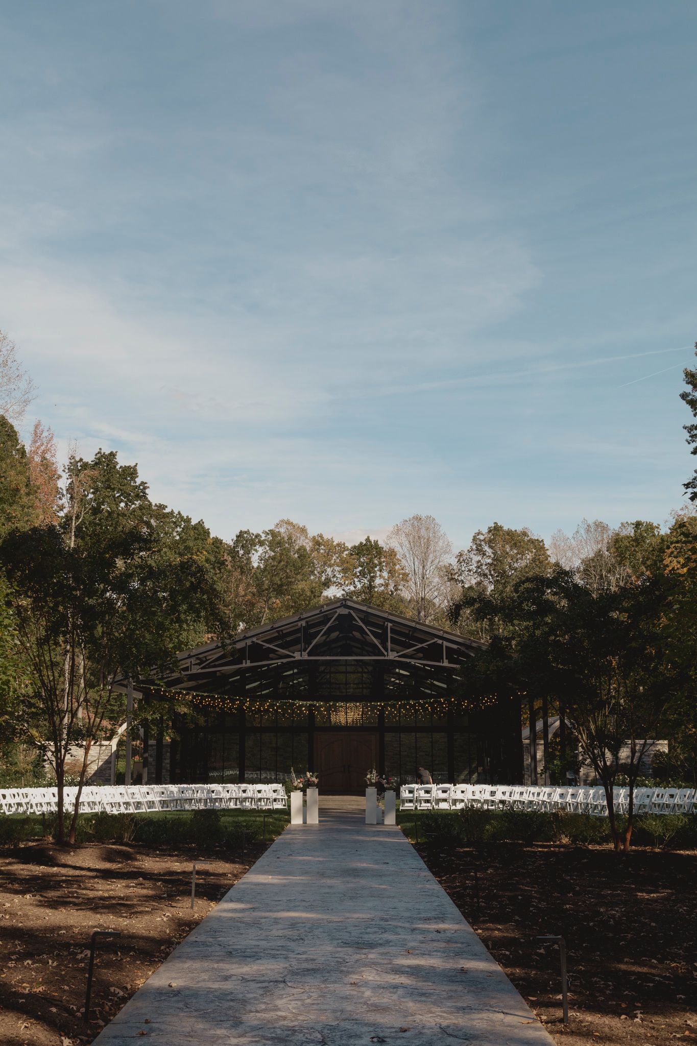 A stone pathway leads to a covered pavilion with white chairs set up for an outdoor wedding ceremony in a wooded setting.