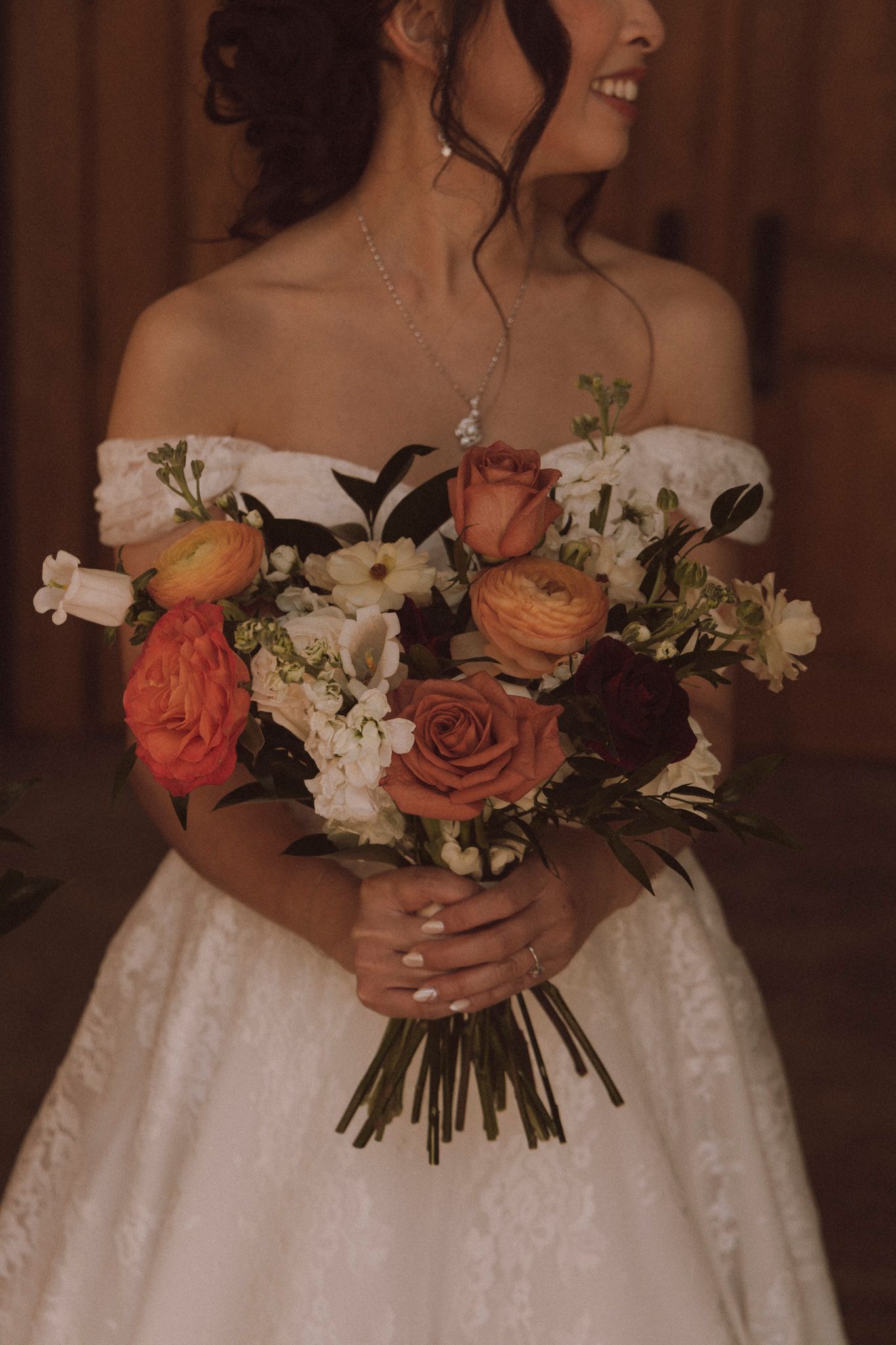 A bride in a white off-shoulder gown holds a lush bouquet of coral and white roses against a dark background.