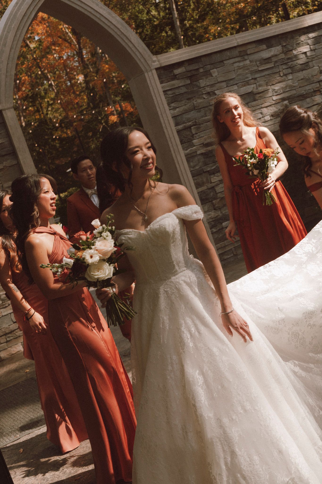 Wedding party poses together in front of stone wall with autumn foliage and arched doorway in background.