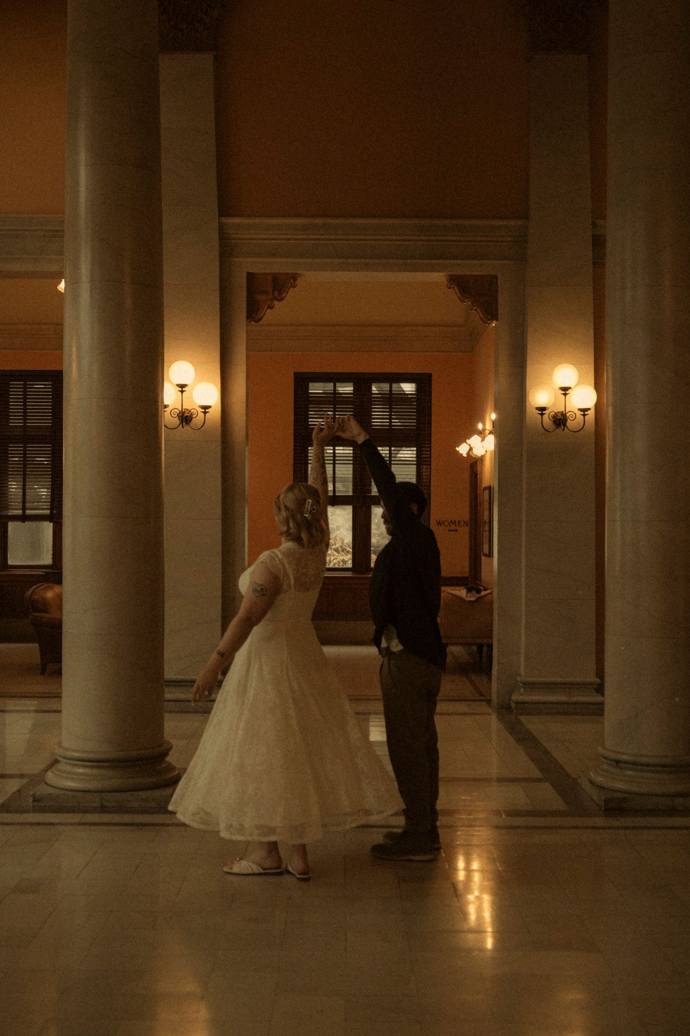 A couple dances in a dimly lit hallway with elegant columns and wall sconces creating a romantic atmosphere.
