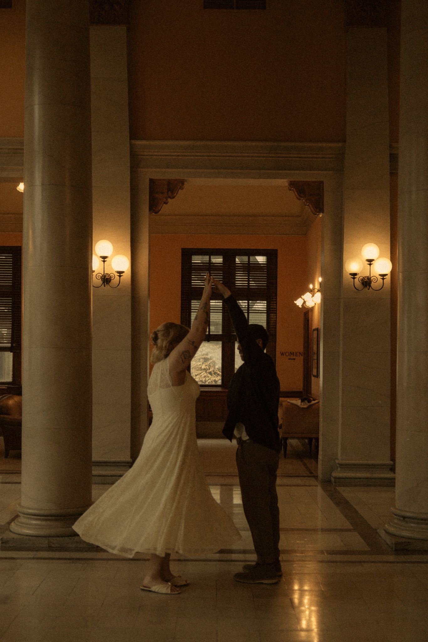 A couple dances together in an elegant hallway with classical columns and warm lighting creating a romantic atmosphere.
