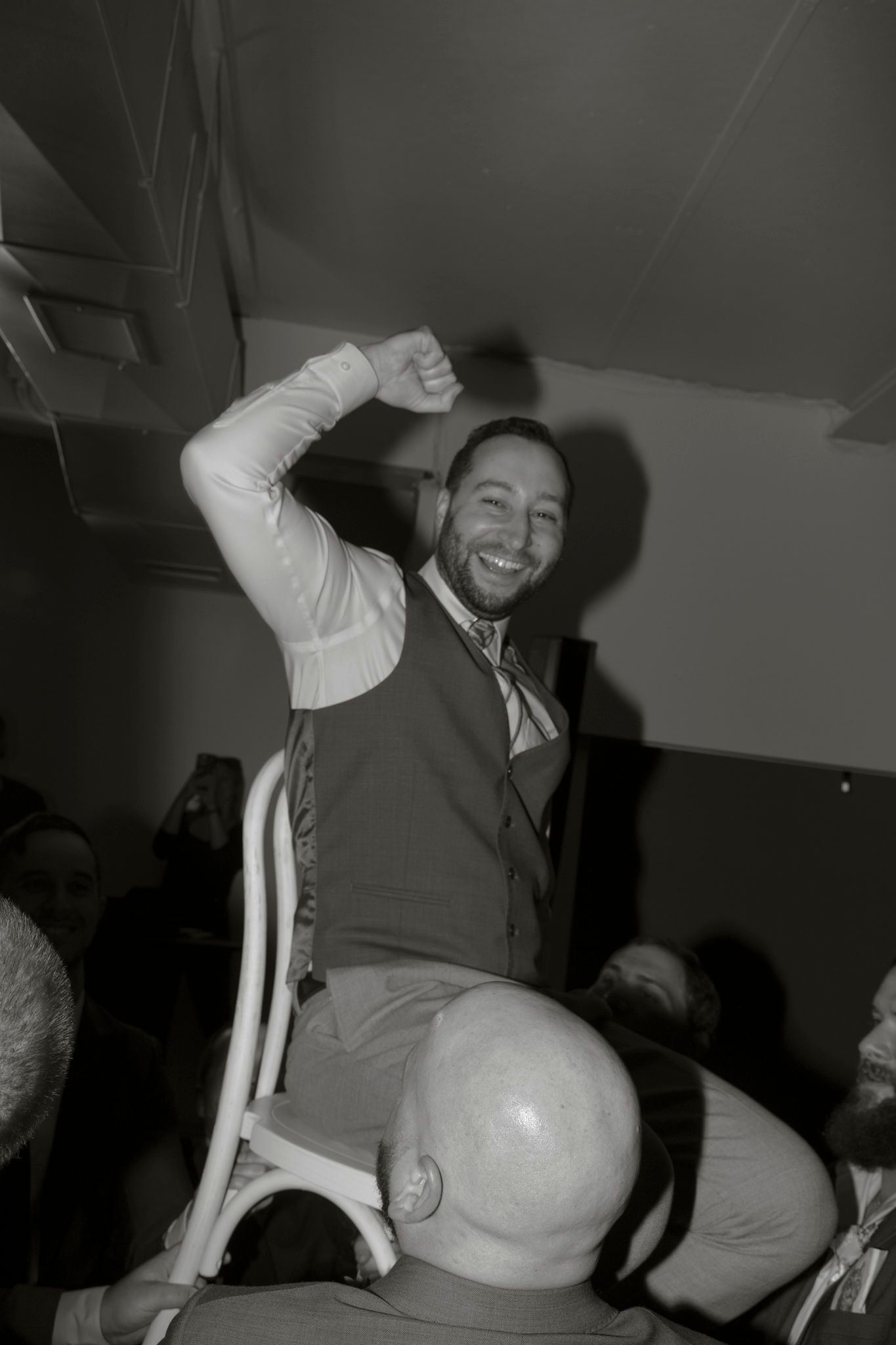 A wedding guest in a vest enthusiastically dances while sitting on a chair.