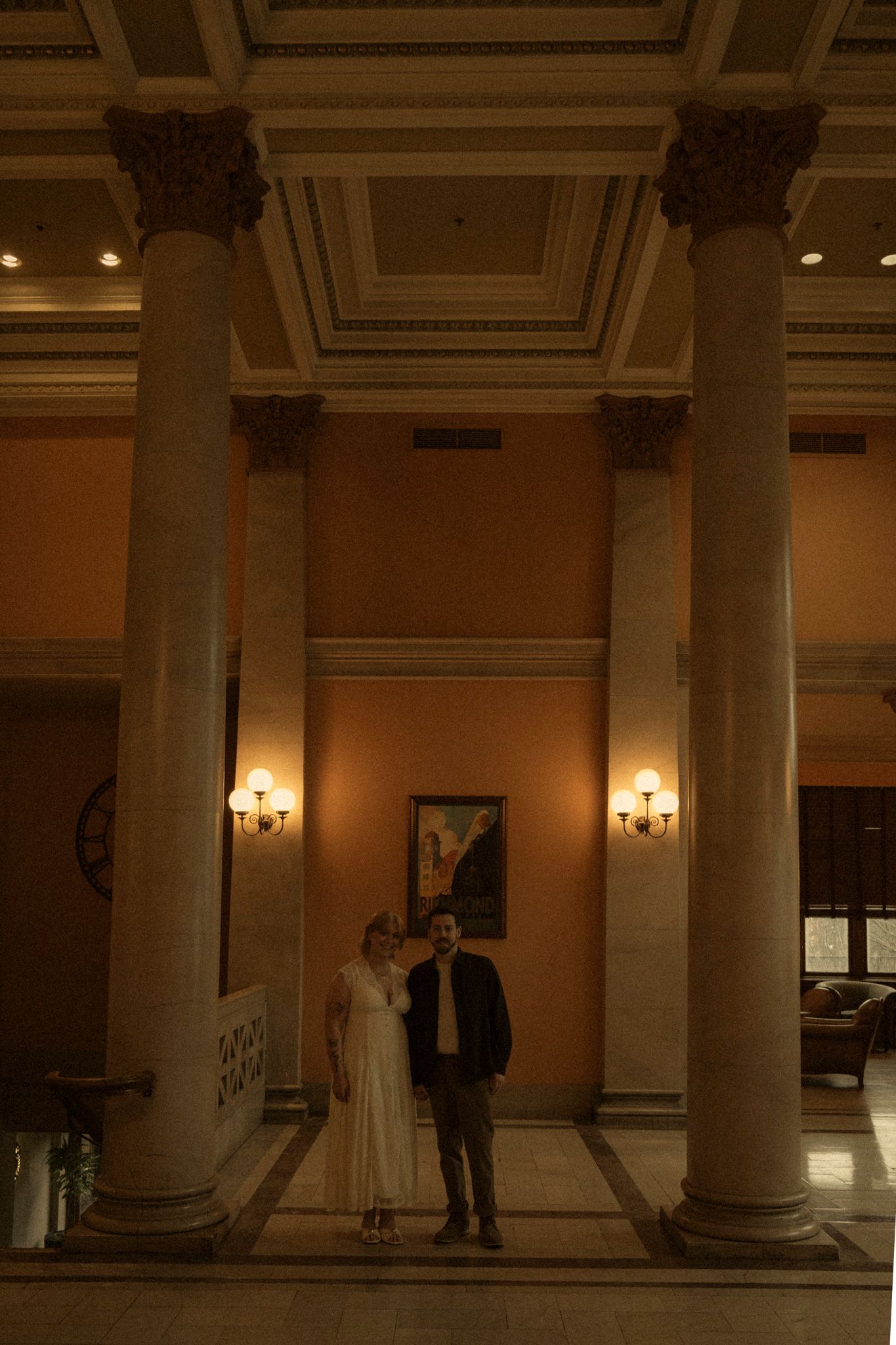 A dimly lit historic interior hallway with classical columns and wall sconces casting warm light on beige walls.