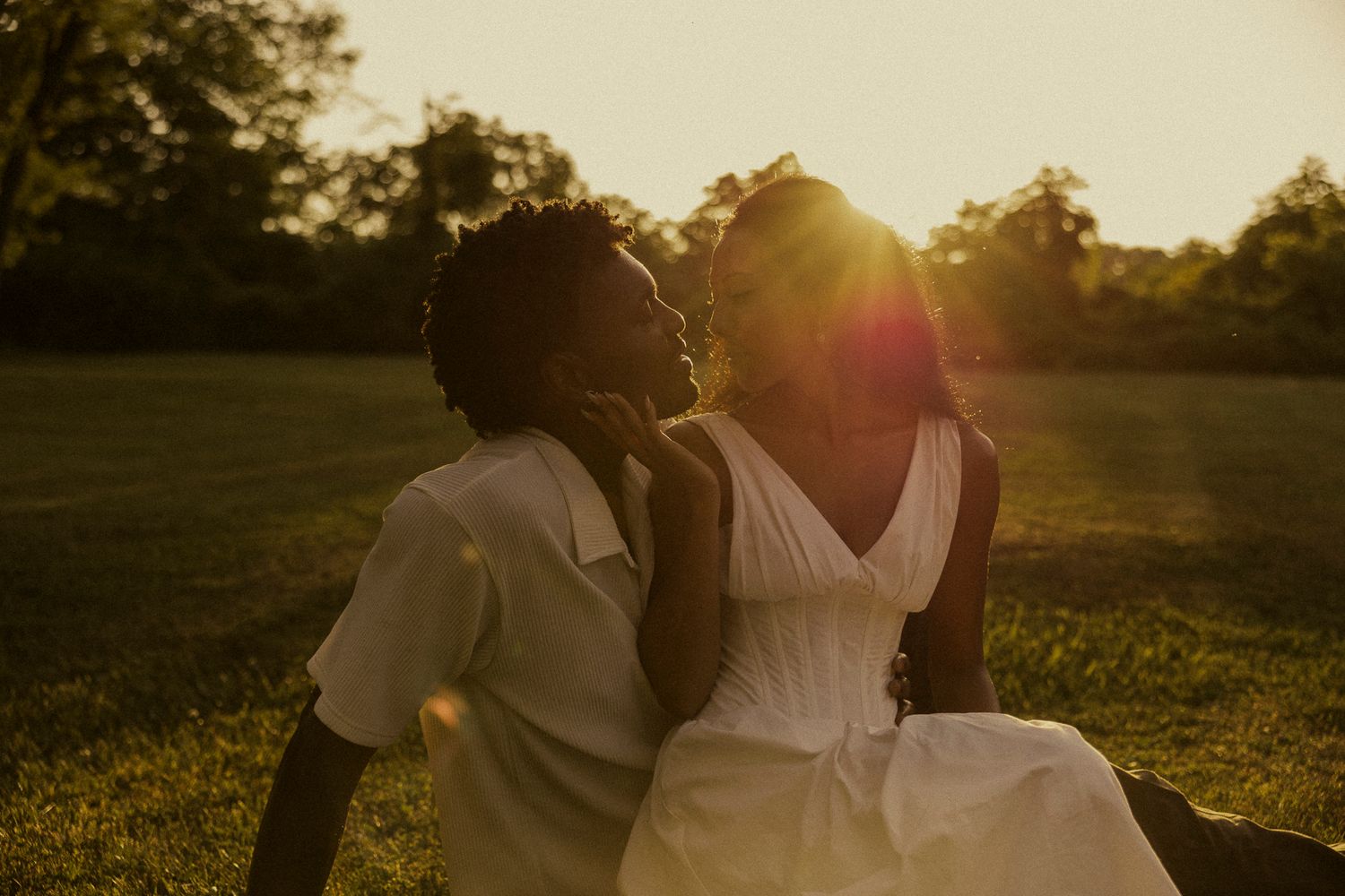 Couple sharing a tender moment at golden hour sunset in a lush green park, bride in white dress.