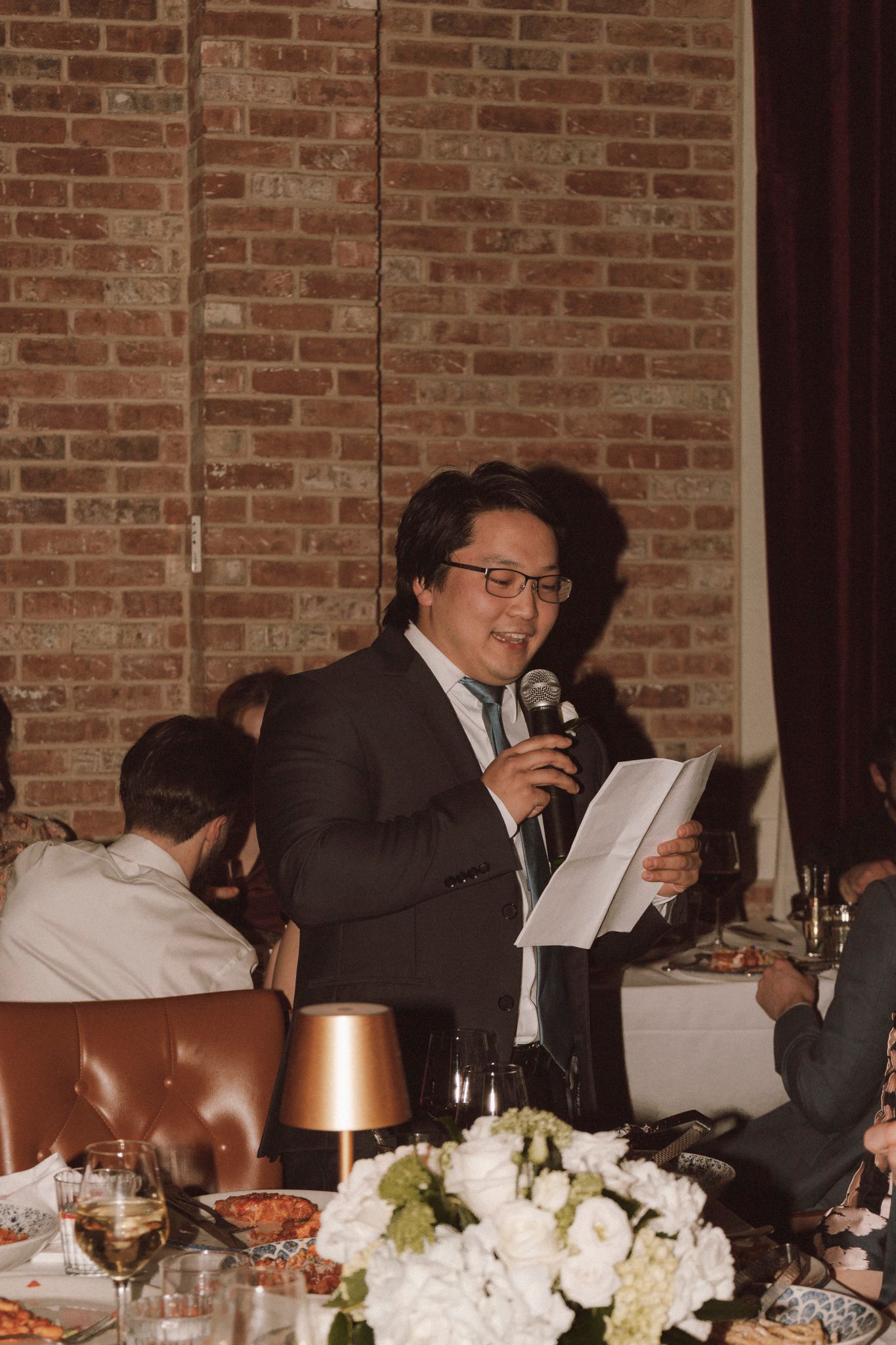 Asian man in suit reads from paper while giving a wedding speech into a microphone at a reception.